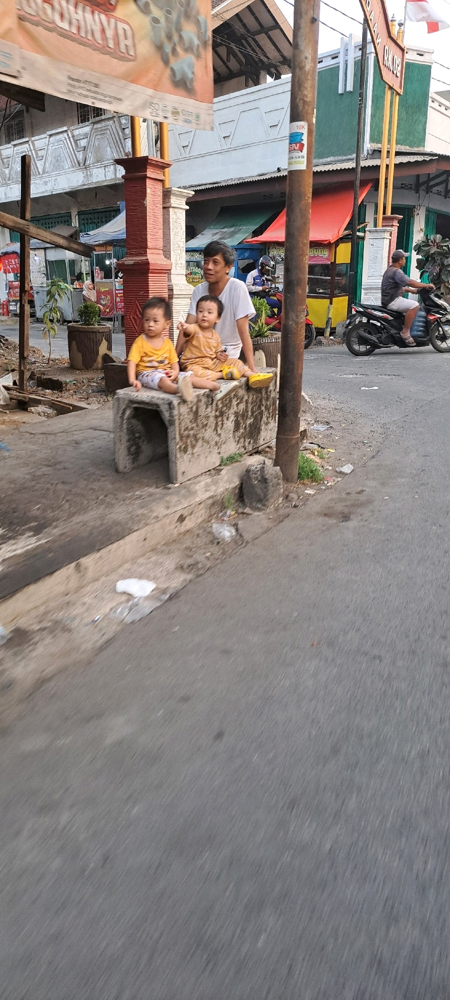 The image captures a street scene in Petungasri, Indonesia. In the foreground, two young children are seated on a concrete structure, one pointing to the left. Behind them, an adult male is standing. The children are dressed in casual clothing. The environment includes a paved road, a sidewalk or curb area with some debris, and buildings in the background. A motorcycle is visible on the right side of the image, with a rider wearing a helmet. A street pole with some stickers is also present. The overall scene appears to be an outdoor, everyday setting.