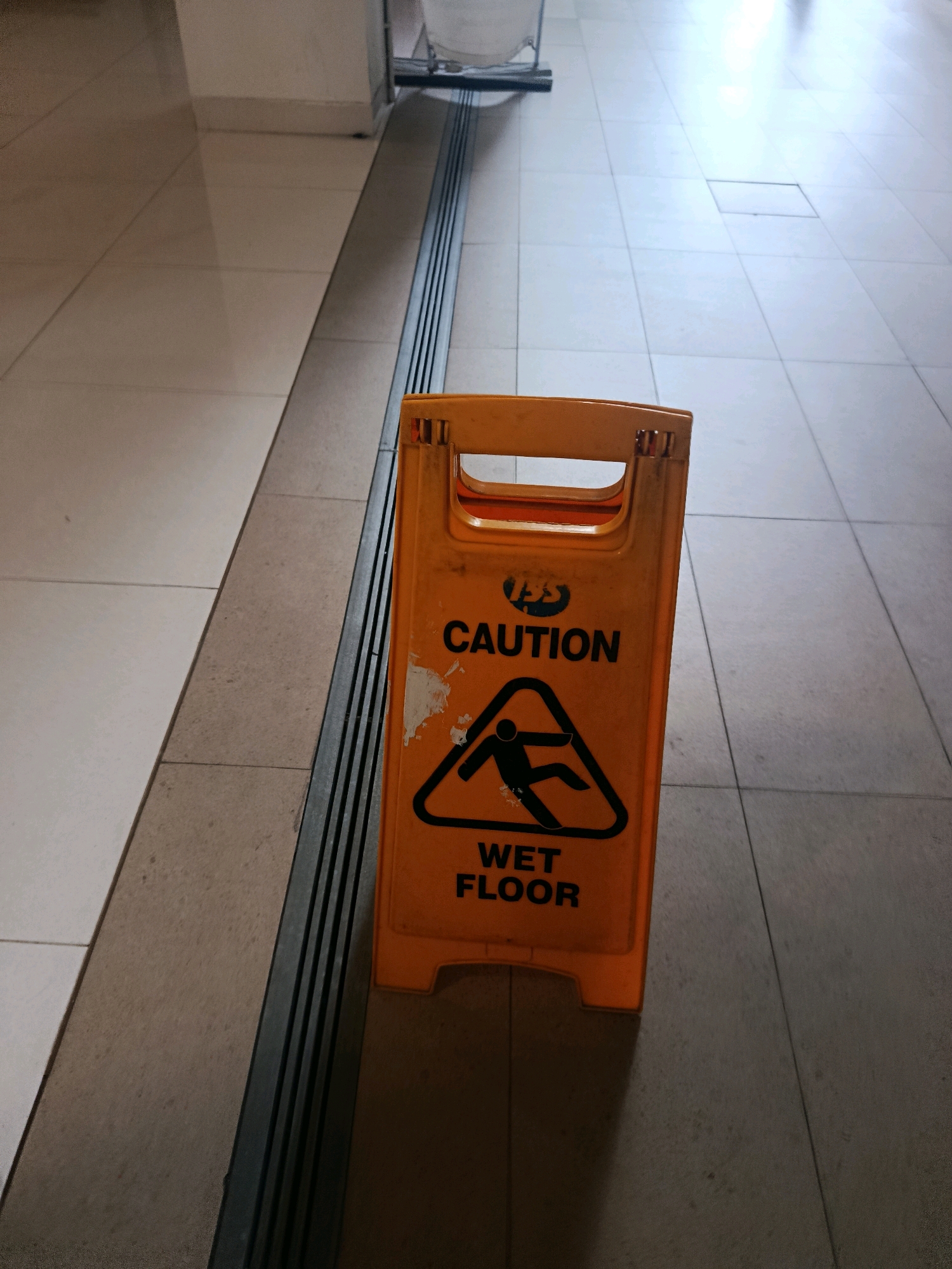 A yellow plastic "Caution Wet Floor" sign stands upright on a tiled floor. The sign has a black symbol of a person slipping and the words "CAUTION" and "WET FLOOR" printed in black. The floor appears to be a light-colored tile, with a dark, ribbed strip running along the left side of the image. The background is out of focus, showing a dimly lit indoor environment. The scene is located in a public space, likely a hallway or lobby, in South Jakarta, Indonesia.