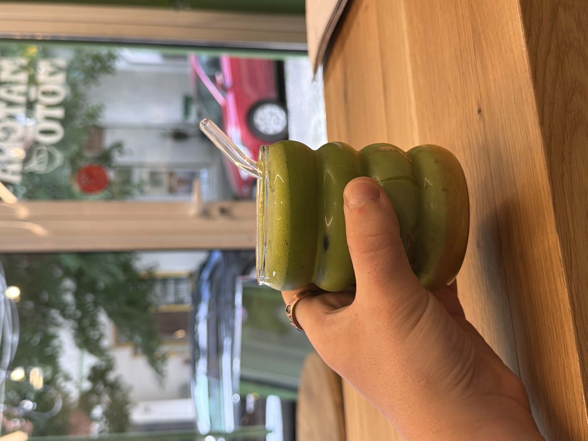 A right-handed person is holding a cup with a green matcha-like beverage and a clear straw. A ring is visible on the person's left ring finger. The cup has a distinct, rounded segmented design. The cup and hand are positioned in front of a blurred window. The window looks out onto a street with trees and a building with white signage that reads "ROTAM OTO". A red car is also visible through the window. The scene is in Sofia, Bulgaria. The hand is held in front of a light brown, vertical wooden surface.