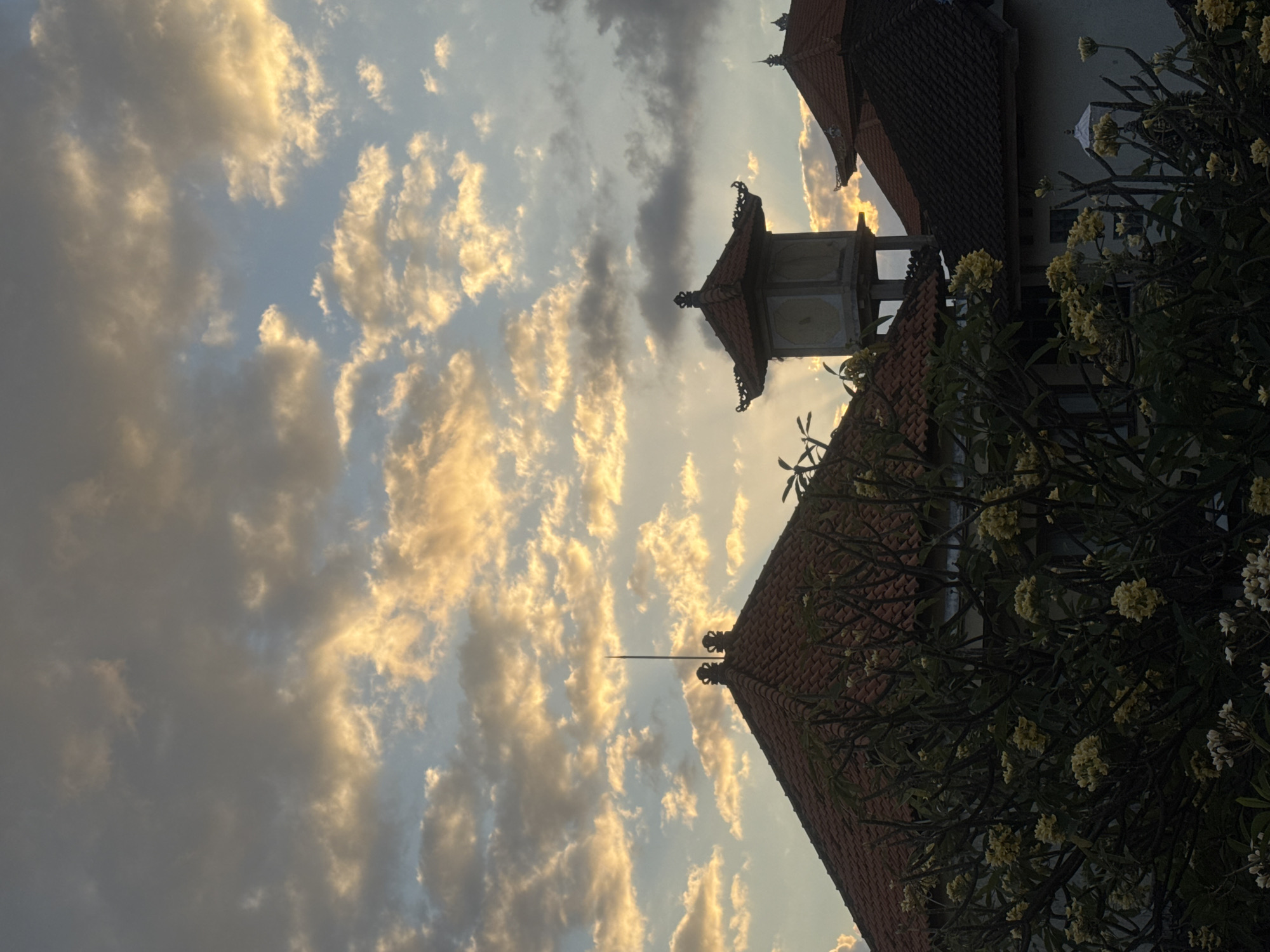 The image shows a view of the sky over Kuta, Indonesia, at sunset. The sky is filled with dramatic clouds, some of which are illuminated by the setting sun, casting a warm, golden glow. In the foreground, several traditional Balinese buildings with red-tiled roofs are visible. One building has an ornate tower with a pagoda-like roof. Lush green trees with yellow flowers are in front of the buildings, partially obscuring them. The scene suggests a peaceful and serene atmosphere, typical of a tropical sunset.