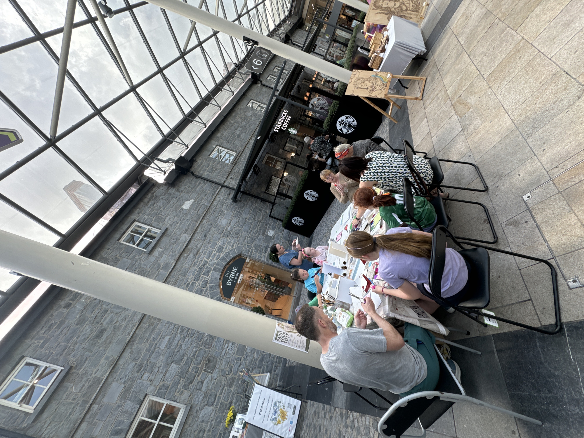 The image depicts an outdoor art workshop event taking place in a covered walkway, likely in Kilkenny, Ireland, given the presence of a "BYRNE EYECARE" shop, a common local business. Several people, including adults and children, are seated around tables, engaged in various craft activities. A man in a grey t-shirt is seen cutting paper with scissors, while a young girl in a purple t-shirt works on a project. Another woman in a green and white patterned top is also participating. In the background, a Starbucks Coffee shop is visible, suggesting an urban commercial area. Easels displaying artwork are set up nearby, with one featuring a portrait. The walkway features a glass and metal roof structure, and the buildings are constructed of grey stone. The ground is paved with stone tiles. The overall atmosphere appears to be casual and community-oriented, focused on creative engagement.