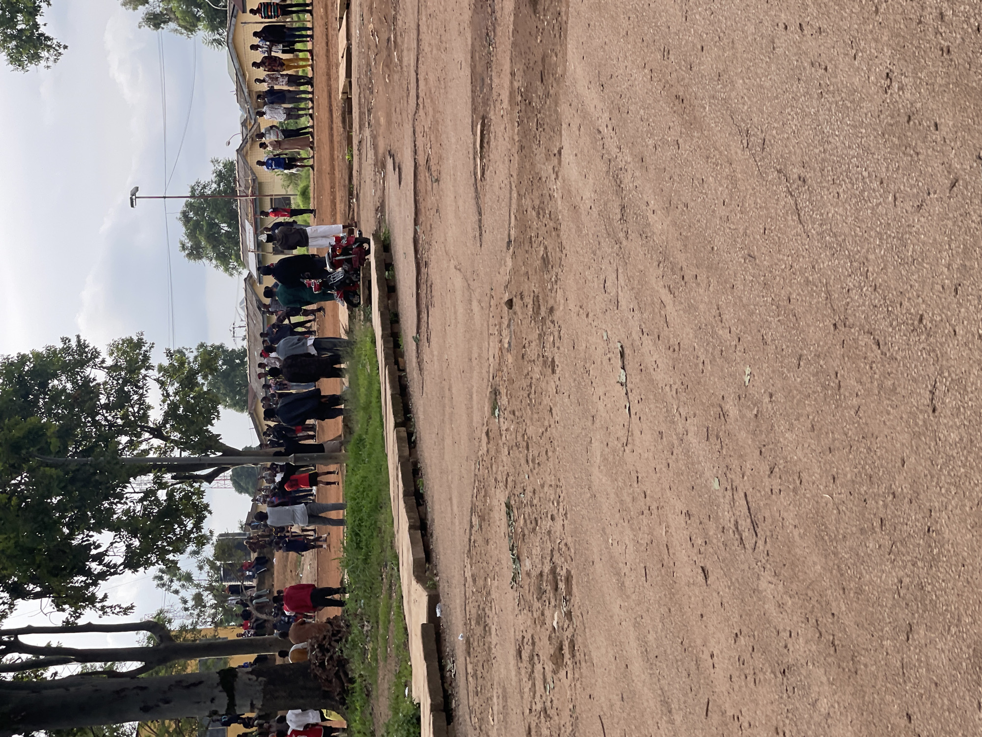 The image depicts an outdoor scene in what is identified as Rafin Par, Nigeria, with a group of approximately 30-40 people gathered. The ground is a reddish-brown dirt, indicating a potentially arid or dusty environment. A raised concrete pathway is visible in the mid-ground, appearing somewhat dislodged in sections. Several trees are present, with lush green foliage, suggesting a moderate climate. In the background, a row of single-story buildings with yellow walls and corrugated roofs are visible. A red motorcycle is parked in the middle ground, facing right. The people are dispersed across the scene, with some standing in groups and others appearing to be in conversation. Their clothing styles are varied, suggesting a mix of casual and possibly work attire. The sky is overcast with visible clouds.
