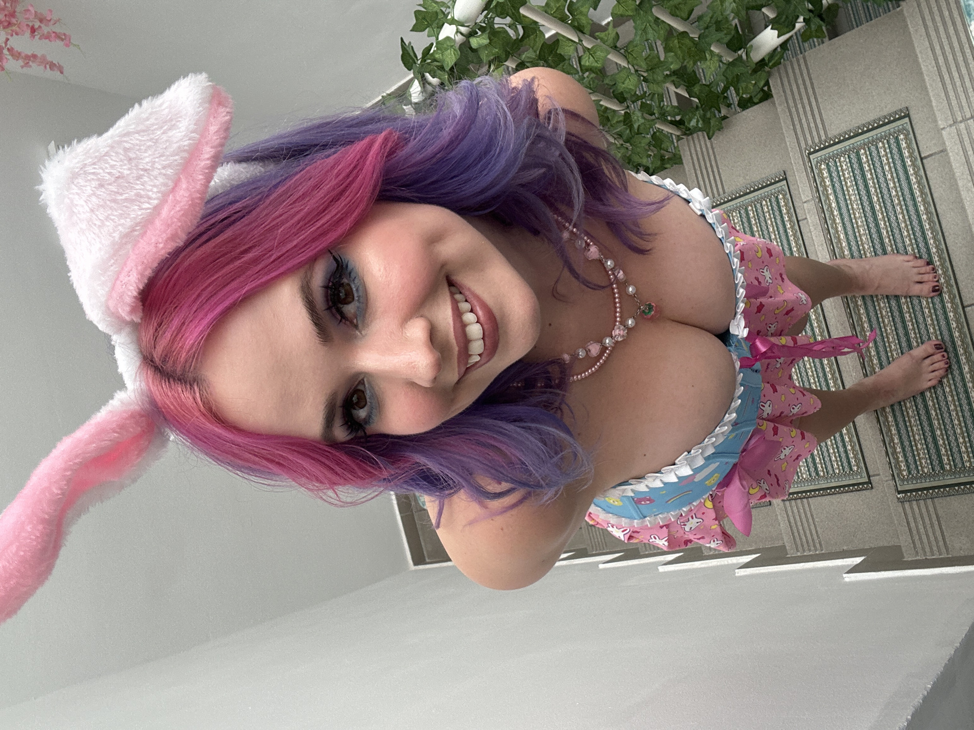 A young woman with purple and pink hair is wearing a pink and white bunny ears headband, a pink and blue dress, and a pearl necklace. She is smiling and looking up at the camera. The background appears to be a staircase with a green plant on top. The location is likely an indoor setting. This photo doesn't provide definitive clues to identify a specific place within the city of Zagreb, Croatia.