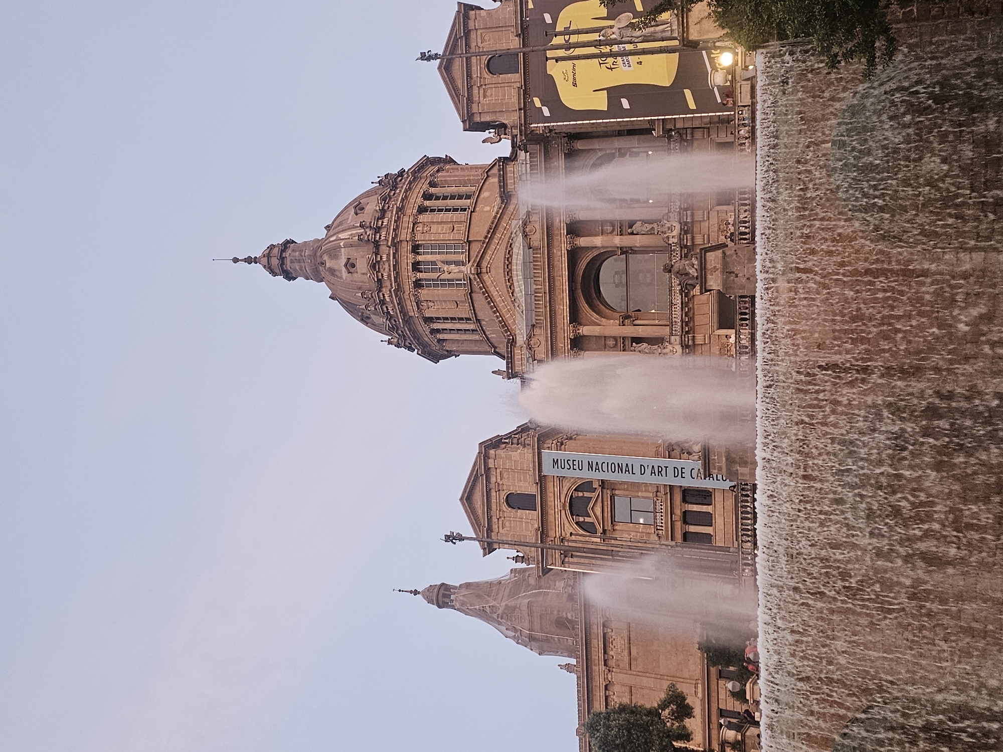 The image shows a frontal view of the Palau Nacional, also known as the National Art Museum of Catalonia, in Barcelona, Spain. A large tiered fountain is in the foreground, with water cascading down its levels. Behind the fountain, the facade of the Palau Nacional is visible, characterized by its grand architecture, domes, and columns. A banner hangs from one of the buildings, displaying text in Catalan. To the right, a large yellow banner with an image of a cyclist and text related to the Tour de France is also displayed. Several people are seen in the background, some standing near the fountain and others on balconies of the building. The sky above is clear and pale, suggesting it is either early morning or late evening.