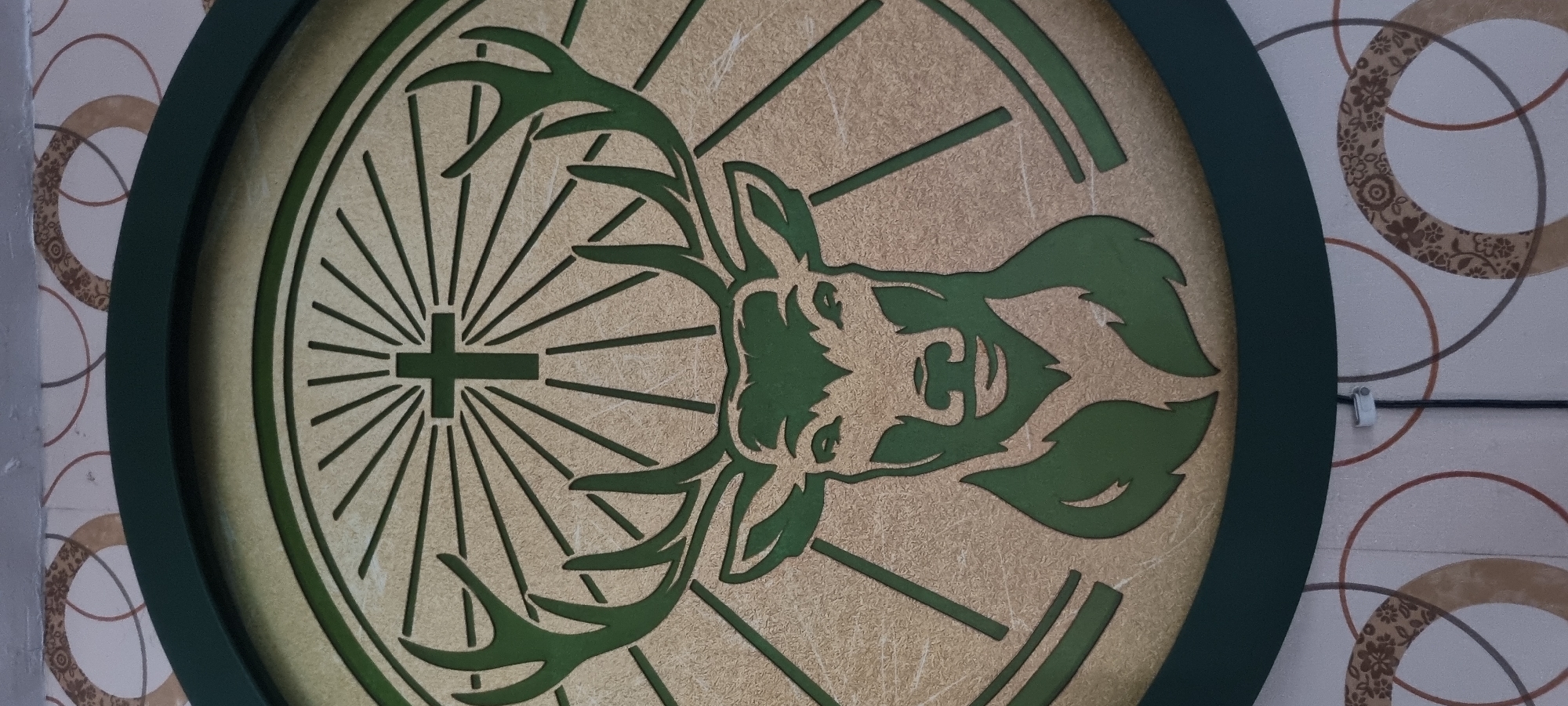 The image shows a circular Jägermeister logo hanging on a wall. The logo features a green deer head with antlers inside a wheel, with a Christian cross positioned above the deer's head. The background of the logo is a textured gold color, and the outer ring is dark green. The wall behind the logo has a patterned wallpaper with circles. The location is unknown; there is no information to indicate the scene is in Alasia, Nigeria.