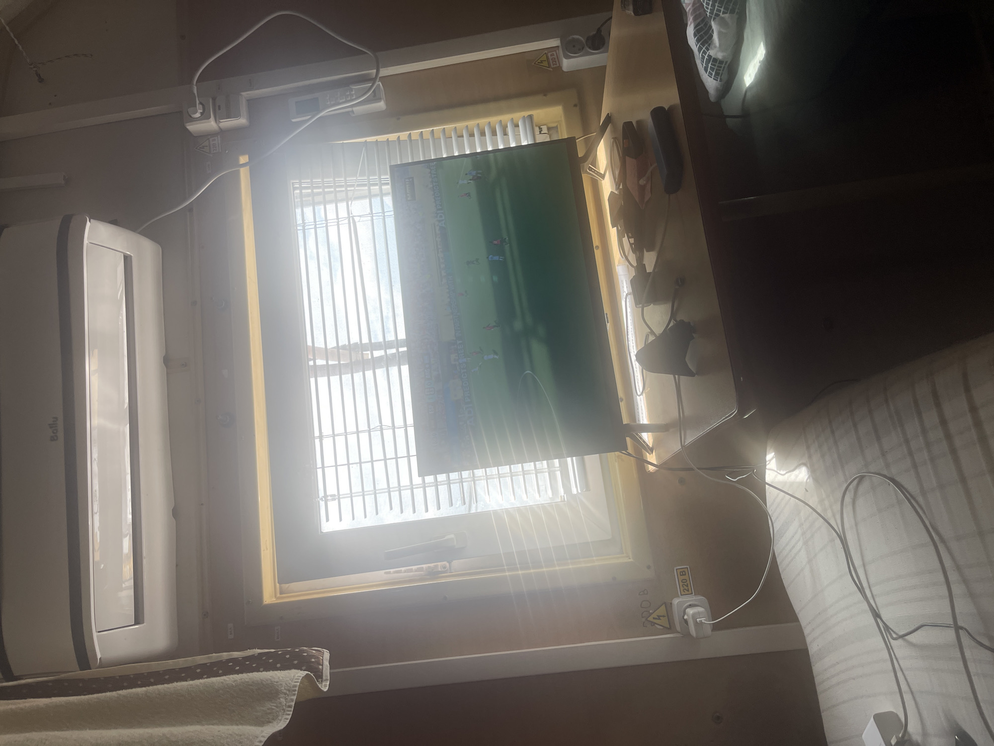 The image depicts an indoor scene, likely a small room or living space, with bright sunlight streaming through a closed window covered by horizontal blinds. A modern flat-screen television is mounted on a stand on a wooden desk, displaying a soccer match. On the desk, there are various electronic accessories, including chargers, cables, and a dark-colored rectangular object, possibly a power bank or external hard drive. To the left of the desk, a white air conditioning unit is mounted high on the wall. A towel with a polka dot pattern hangs from the left side of the frame. Electrical outlets and warning labels are visible on the wall to the right of the window. The foreground shows a portion of a bed with a striped mattress protector. The overall impression is of a functional, albeit somewhat cluttered, personal space. The location is stated to be Tatishchevo, Russia.