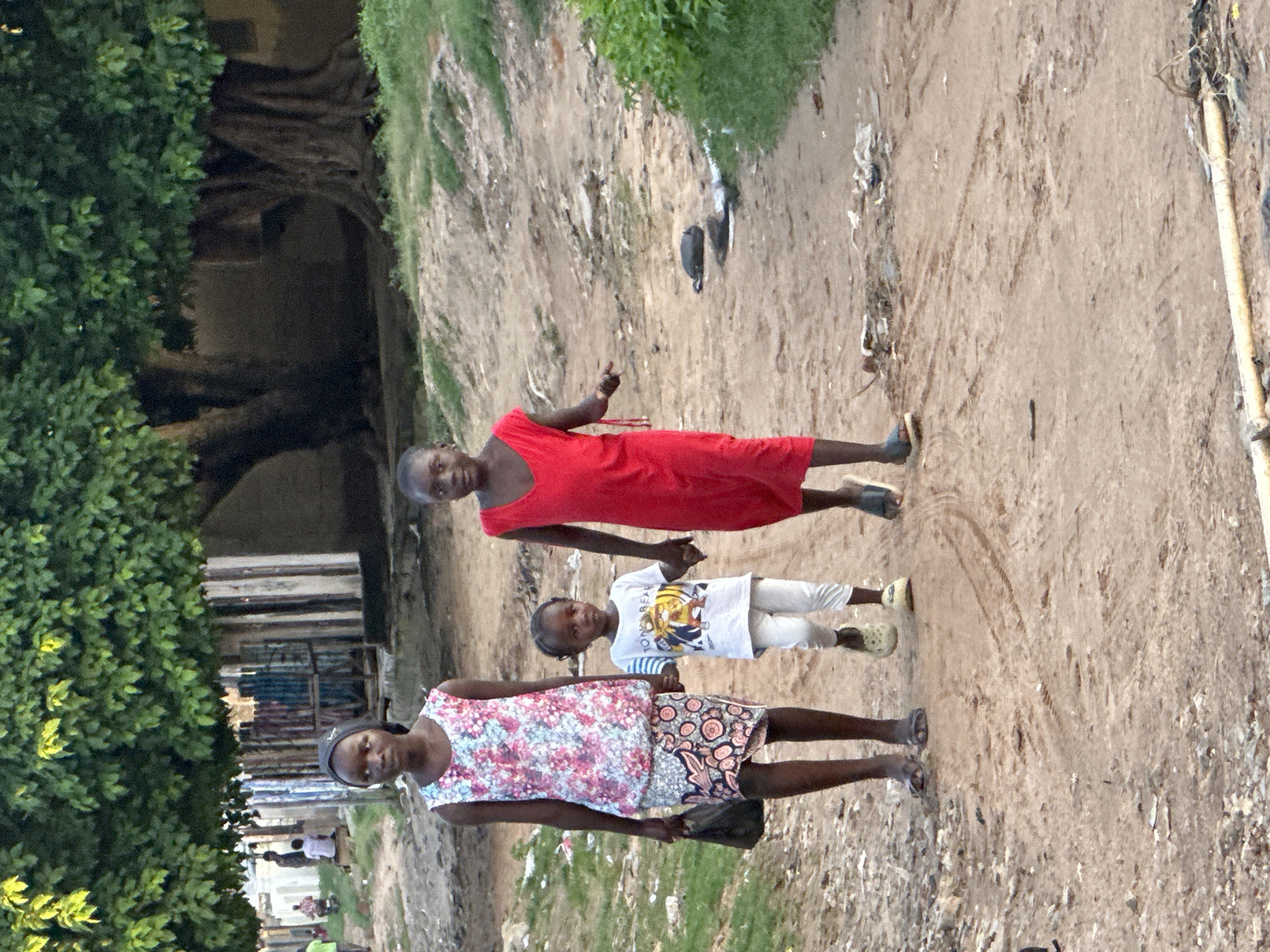 Three children are walking on a dirt road. The child on the left is wearing a floral sleeveless dress and a headscarf. She is carrying a black bag in her left hand and holding the hand of the child in the middle. The child in the middle is wearing a white t-shirt with a graphic print and white pants. The child on the right is wearing a red sleeveless dress. They are walking towards the right side of the image. In the background, there are trees and structures, suggesting an outdoor, possibly rural or informal urban setting. The ground is sandy and uneven, with some debris visible.