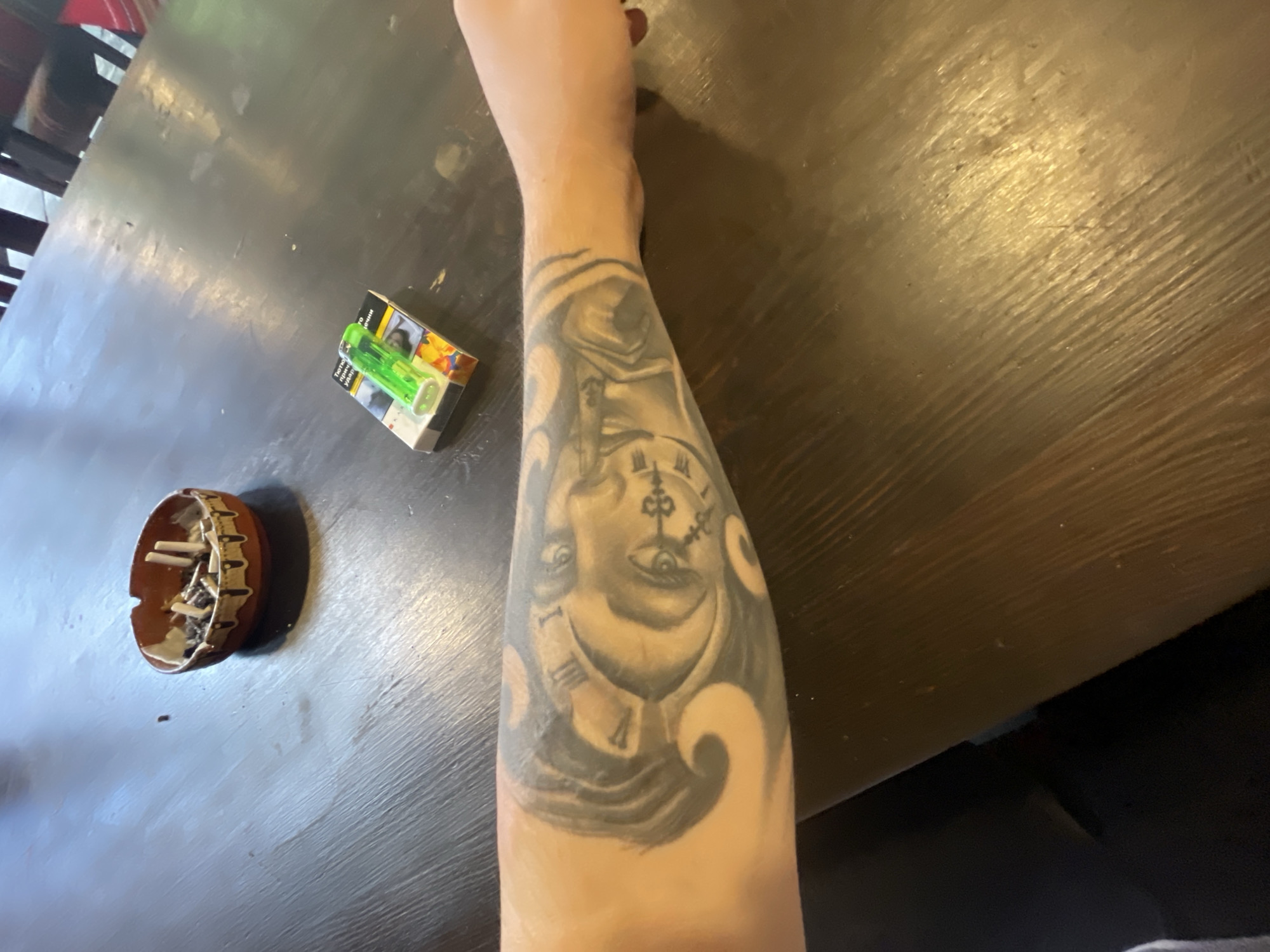 In an indoor setting, a person's tattooed forearm rests on a dark wooden table. The tattoo depicts a clock face with Roman numerals and a human face integrated into the design. To the right of the forearm are a pack of cigarettes and a green lighter. Further to the left, an ashtray filled with cigarette butts sits on the table. The table surface exhibits a wood grain texture. The background is out of focus, showing glimpses of chairs and possibly a bar area, suggesting the location is a bar or pub.