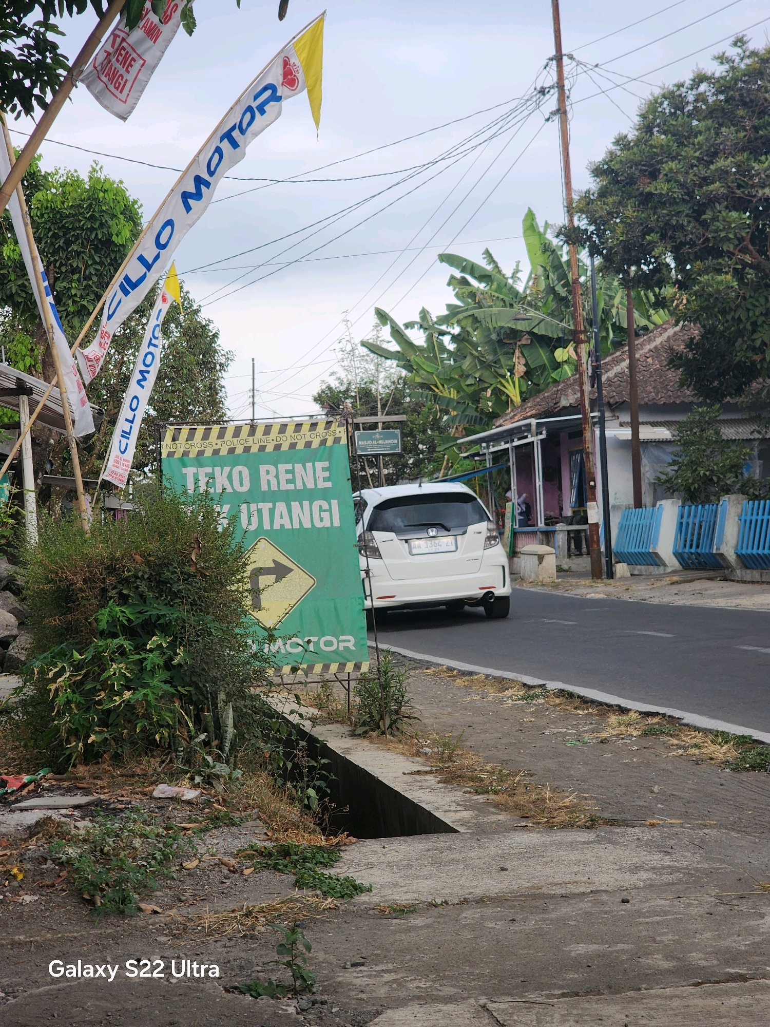 In Salaman, Indonesia, a white Honda Fit is parked on the right side of a road, facing away from the viewer. A green billboard with white text and a yellow directional arrow sign is positioned to the left of the parked car. The billboard reads "TEKO RENE UTANGI MOTOR". Above this, several flags on poles display text, including "CILLO MOTOR". The scene is outdoors with a cloudy sky. There are trees and a building visible in the background. The foreground shows a grassy area and a concrete structure, possibly a drainage ditch. The overall impression is of a street scene in a semi-urban or rural area.