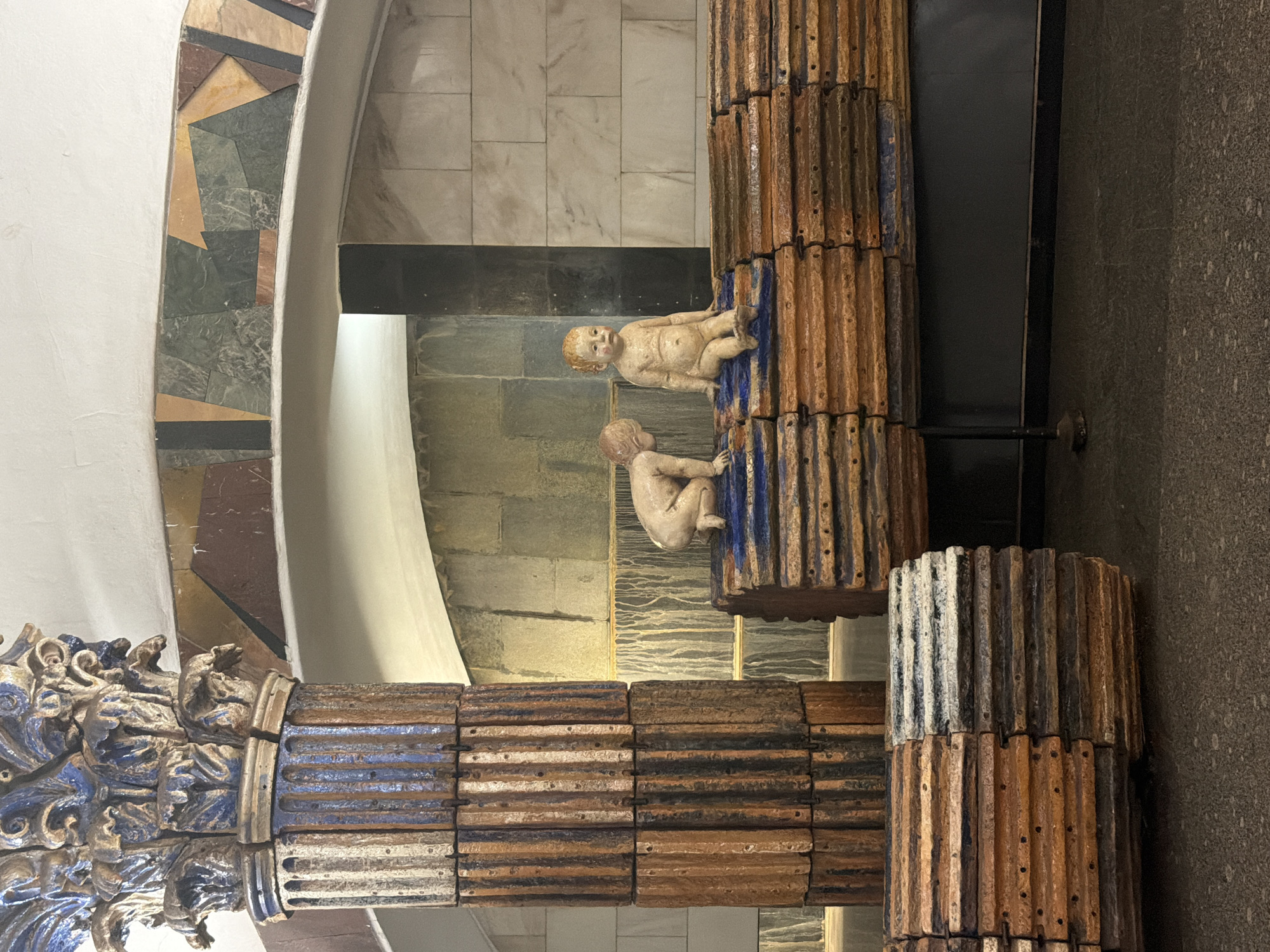 The image depicts an interior scene within **Partizanskaya Metro Station in Moscow, Russia**, characterized by unique architectural and sculptural elements.
In the foreground to the left, a large, cylindrical column rises, constructed from vertically stacked, rough-textured rectangular blocks predominantly in shades of reddish-brown and dark blue-grey. The column's capital is ornate, featuring intricate, almost eroded-looking blue and gold/brown acanthus leaf motifs.
Extending horizontally across the lower midground is a multi-tiered structure, resembling a low bench or platform, made of similar rough-textured, stacked rectangular blocks in reddish-brown with blue accents. This structure is supported by slender dark metal legs.
Two light-skinned, naked baby sculptures are positioned on this structure. The left baby is in a crawling or kneeling pose, leaning forward with its head turned to the right. The right baby sits upright on a higher tier, facing forward with its legs tucked. Both sculptures exhibit a matte, aged finish and stylized features.
The background features textured walls composed of varying stone blocks: a pale, variegated grey-brown section behind the sculptures and a lighter, possibly marble-like tiled section to the right. An arched opening frames the upper background, decorated along its curve with a mosaic band of geometrically cut stone pieces in dark green, brown, black, beige, and reddish-brown hues. The ceiling above the arch is white and gently curved. The floor is a dark, speckled, unpolished surface. The overall lighting is diffuse, creating soft shadows and emphasizing material textures.