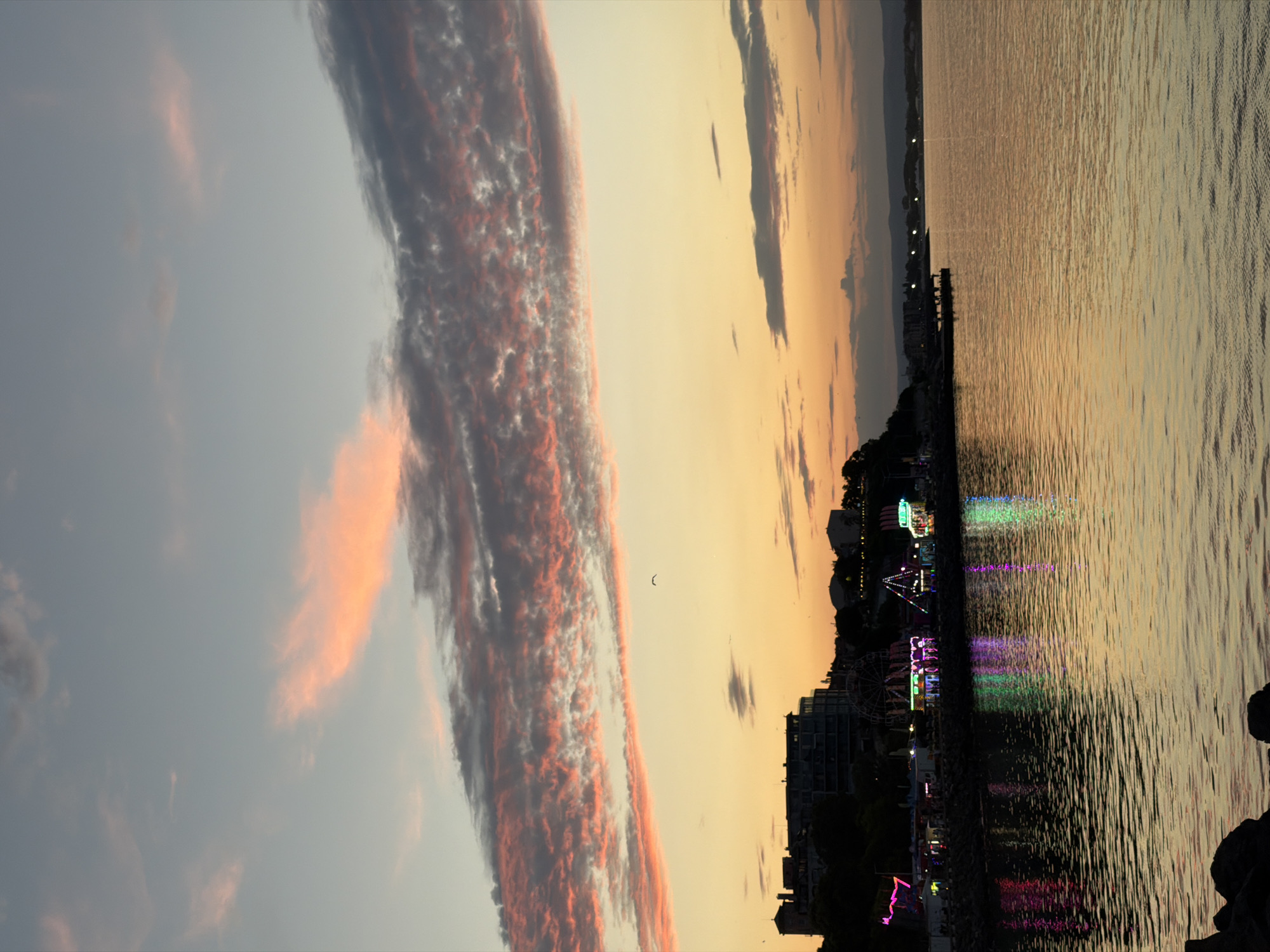 The image captures a twilight scene along the coast of Nesebar, Bulgaria. The sky is painted with hues of pink, orange, and pale blue, with scattered clouds adding texture. Below the sky, a silhouette of buildings lines the coastline, with lights from what appears to be an amusement park or fairground illuminating the scene with vibrant pink, green, and purple reflections on the calm water. The sea stretches towards the horizon, reflecting the sky's colors and the artificial lights from the shore. Dark, rocky formations are visible in the foreground, partially submerged in the water. The overall atmosphere is serene and picturesque, typical of a coastal town at dusk.