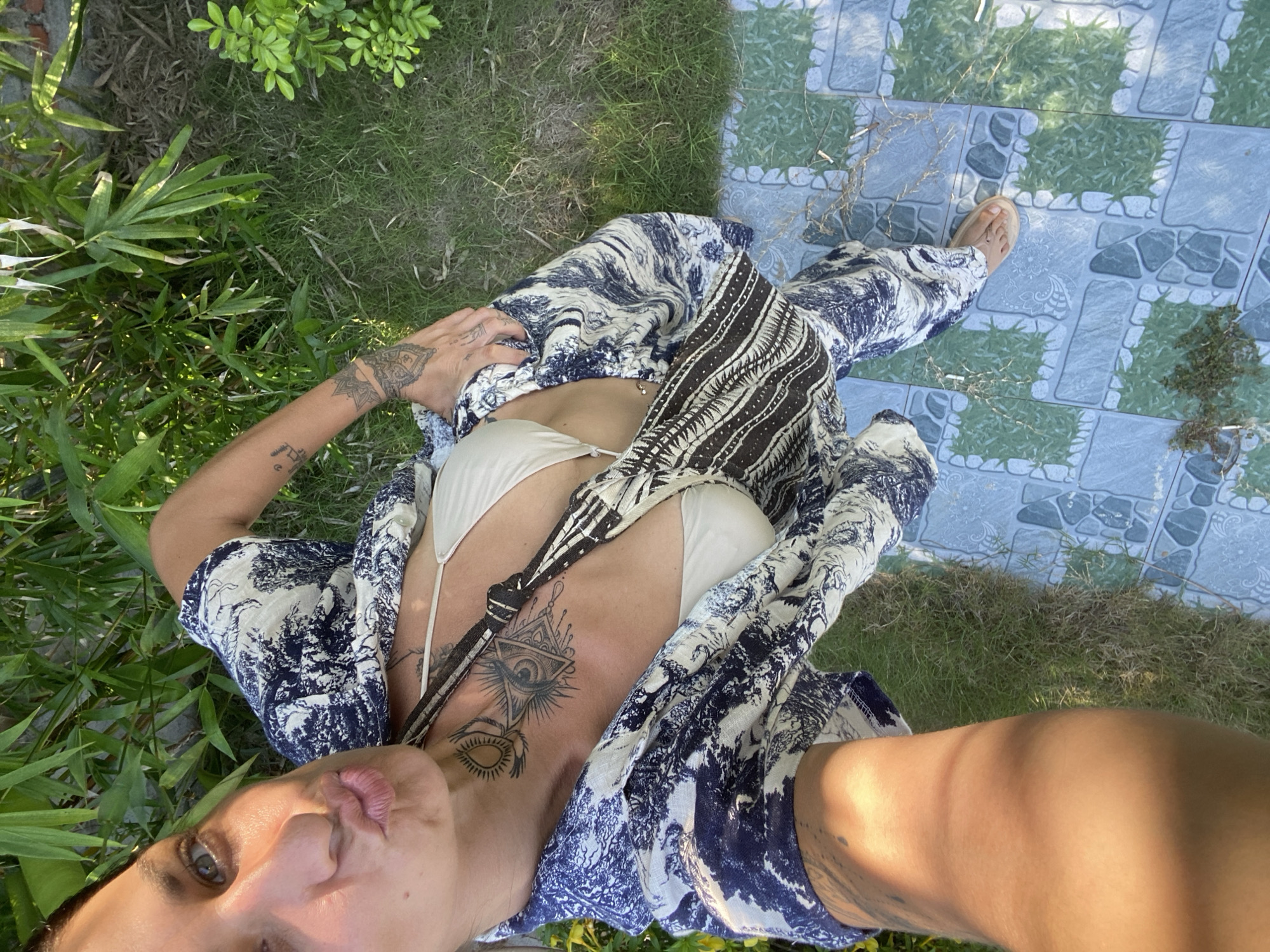 A person is taking a selfie outdoors, posing with puckered lips. They are wearing a blue and white patterned shirt and matching pants, along with a beige bikini top. A patterned crossbody bag is slung over their shoulder. Tattoos are visible on their neck and arm. The person is standing on a tiled walkway that transitions to a grassy area with lush green plants. The scene appears to be a residential or resort area in Mũi Né, Vietnam, with the sunlight casting shadows.