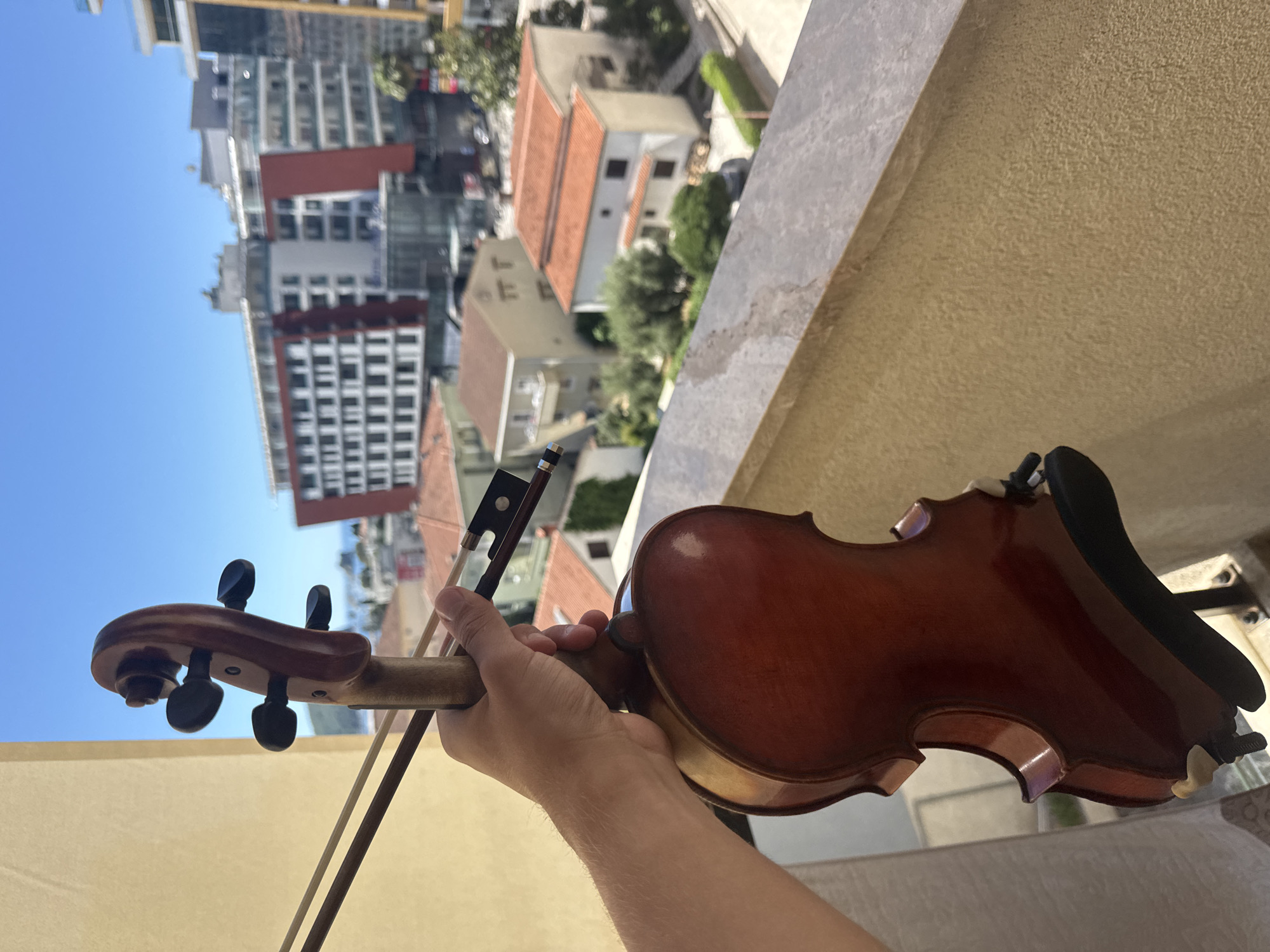 A person's hand holds a violin and bow over a balcony railing, with a clear blue sky and buildings in the background. The buildings appear to be in a coastal urban setting, likely Budva, Montenegro, given the architecture and landscape. The focus is on the violin, which is made of polished wood and has a dark-colored bow resting on it. The hand is holding the neck of the violin, and the bow is positioned to play. The background shows a mix of residential and commercial buildings, some with red-tiled roofs, and palm trees, suggesting a warm climate. The image captures a moment of leisure or practice, with the violin seemingly ready to be played against the backdrop of a vibrant city.