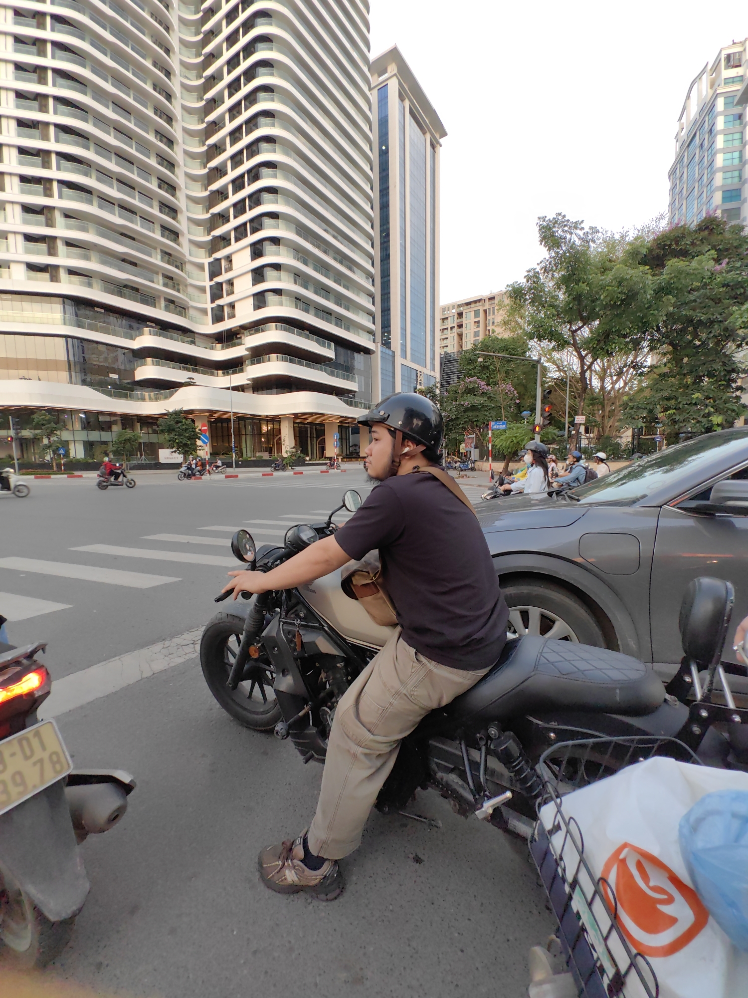 This media file captures a street scene in an urban environment, likely Hà Nội, Vietnam. The central subject is a person seated on a dark-colored motorcycle, stopped at an intersection. The person wears a black t-shirt, khaki pants, and a black helmet, with a strap visible over their right shoulder. Their right hand is on the motorcycle's handlebar, and they are looking forward and slightly to the left.
The motorcycle has a textured black seat and a wire basket on the rear, containing a white shopping bag with an orange logo and a blue plastic bag. In the immediate foreground to the left, the rear wheel and partially visible license plate ("3-01 / 99.78") of another motorcycle are seen. To the right, a gray car is stopped, and further back, other motorcycles and pedestrians are visible at the intersection, with some individuals wearing face masks and helmets. A red traffic light can be seen in the distance.
The background is dominated by modern, tall buildings, one featuring distinctive curved white balconies and large glass windows. Another building with a sleeker, glass-dominant facade is also visible. Green trees line the street, adding natural elements to the urban landscape. The lighting suggests late afternoon or early evening, with clear weather. The street features marked crosswalks. The scene depicts a typical busy city environment with a mix of vehicle traffic and contemporary architecture.