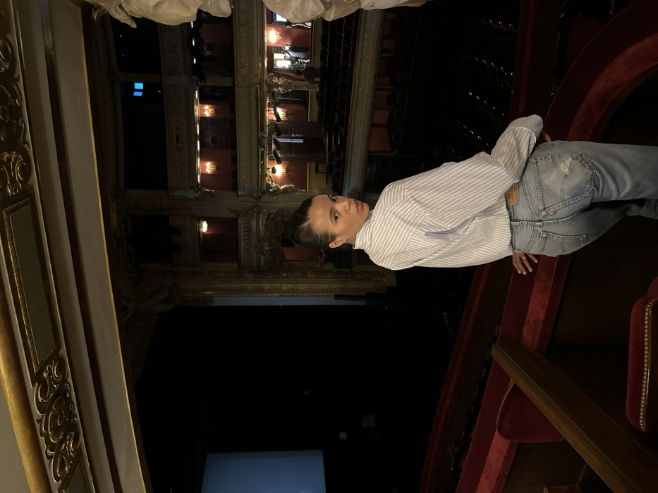 A female individual is captured leaning against a red velvet railing within the ornate interior of a grand theater, likely the Opéra Comique (Salle Favart) or a similar historic venue in Paris, France. The subject, a woman in her 20s or 30s with dark hair pulled back, is positioned in the mid-ground, looking directly over her right shoulder towards the camera. She wears a light blue and white striped collared shirt, untucked, and faded light blue distressed denim jeans with a brown belt. A red Levi's tag is visible on the right rear pocket of her jeans.
The theater's interior features multiple tiers of balconies, extensive gold leaf ornamentation on architectural details and ceilings, and dark red velvet upholstery on railings and seats. The main seating area and stage are largely dark, though a large, light-colored projection screen is visible on the far left side of the stage. Several indistinct figures, appearing to be other patrons or staff, are faintly visible in the upper right background within the tiered boxes. The scene is dimly lit, with the subject prominently illuminated, suggesting a flash or directed light source.