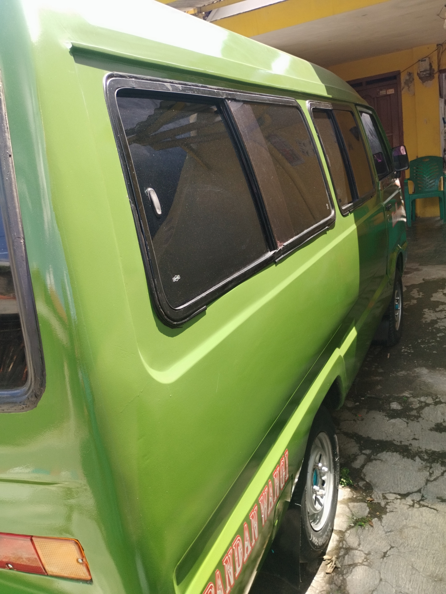 A bright green minivan, likely a Suzuki Carry or similar model, is parked on a concrete surface in front of a yellow building. The van has tinted windows and a red and white decal on its side that reads "ANDAN WANGI" in large letters. The building appears to be a residential or commercial structure, with a yellow facade and a wooden door visible. A green plastic chair is placed to the right of the van. The scene is lit by bright sunlight, casting shadows on the ground. The overall impression is one of a typical street scene in a warm climate, possibly in the city of Surabaya, Indonesia.