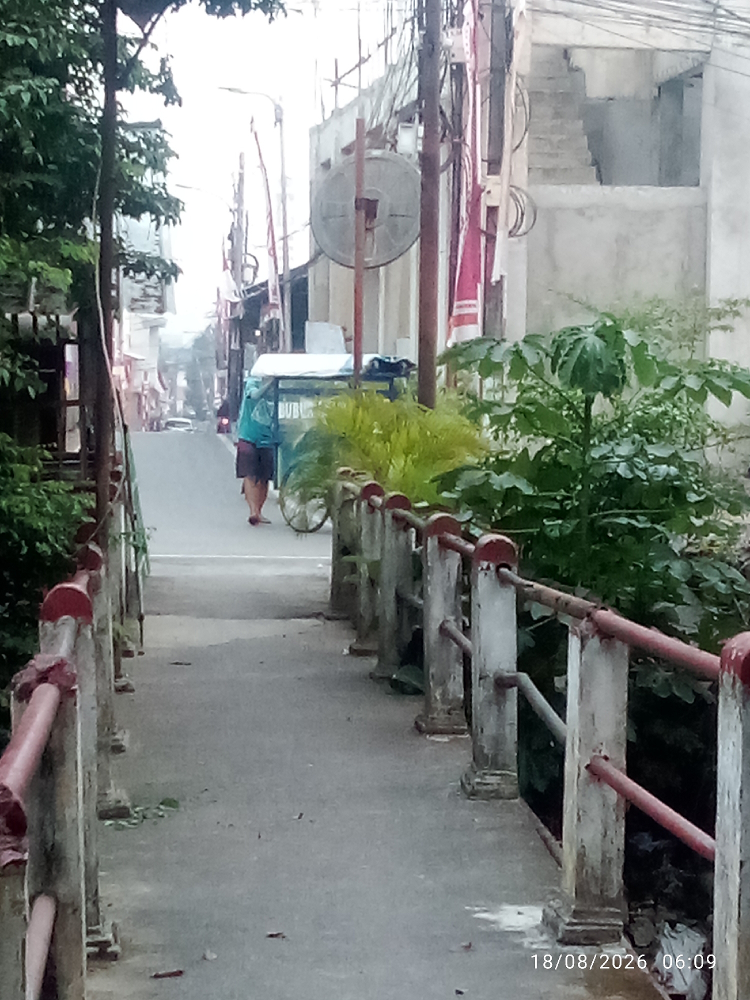 A person wearing a turquoise shirt and dark shorts walks away from the camera on a road that is lined with trees and buildings. The person is pushing a cart with a white canopy. The cart has "BUBBLE" printed on its side. The road is wet, and there is a bridge in the foreground with red railings. The date and time stamp "18/08/2026 06:09" is visible in the bottom right corner. This scene is likely in East Jakarta, Indonesia, given the architecture and presence of a street vendor.