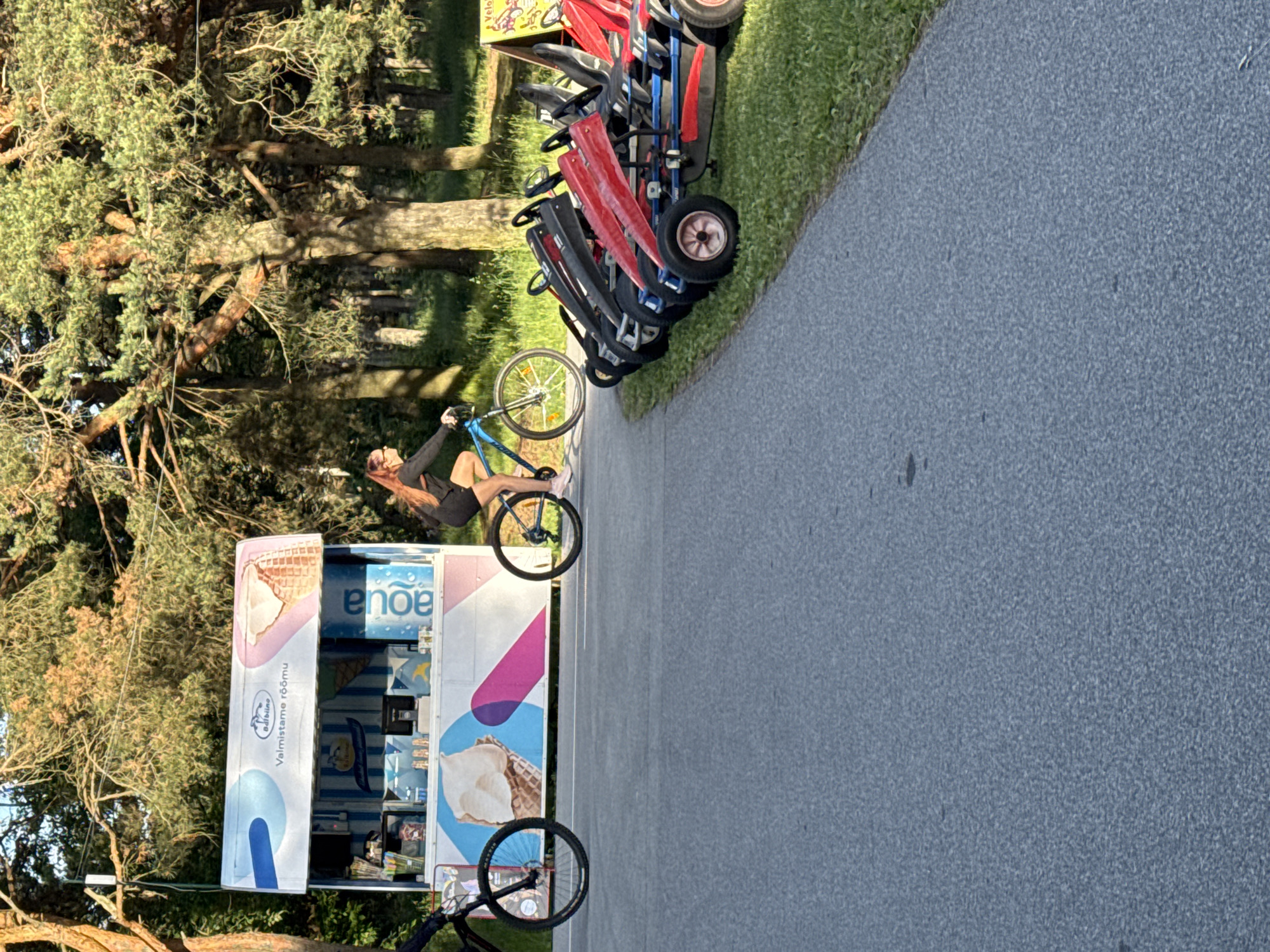 The image depicts a sunny outdoor scene, likely in a park or recreational area in Tallinn, Estonia, given the presence of an ice cream cart and pedal go-karts. The primary subject is a young woman with reddish hair, wearing a dark top and shorts, riding a blue bicycle on a paved path. She is in motion, appearing to cycle away from the viewer towards the right side of the frame.
To the left of the path is a brightly colored ice cream cart with an awning displaying imagery of ice cream cones and the text "Valmistame rõõmu" (We make joy) and "aQua". The cart appears open for business, with items visible on display. To the right of the path, several pedal go-karts are parked in a row on a grassy area. They are a mix of red and black colors, with blue accents.
In the background, a dense stand of pine trees provides shade and a natural environment. The ground is a mix of paved path and green grass. The lighting suggests it is daytime, with sunlight filtering through the trees. The overall atmosphere is one of leisure and recreation.