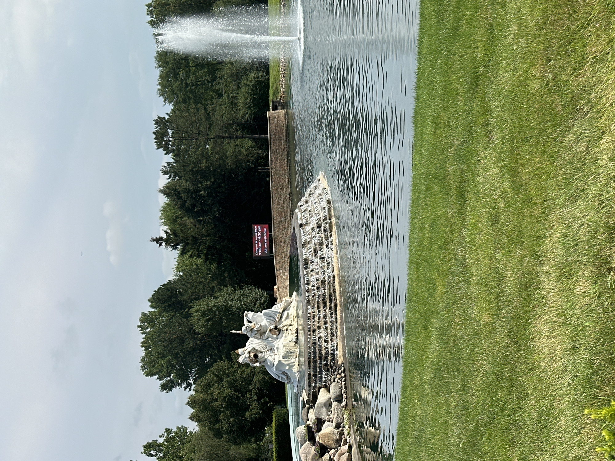 The image depicts a serene outdoor scene with a large pond and a cascading fountain as the central focus. In the background, lush green trees form a dense border, with a clear, slightly overcast sky above. To the right of the fountain, a tall plume of water sprays upwards, its droplets catching the light. The pond's surface is calm, reflecting the surrounding greenery and the sky.
On the left side of the pond, a sculpted white statue sits atop a tiered rock formation from which water flows down into the pond. The statue appears to depict several figures in a dynamic pose. Further to the right, a red digital sign is visible, displaying text that reads: "Campus is open from 9:00 AM - 5:30 PM All are welcome!".
In the foreground, a neatly mowed lawn extends towards the edge of the pond, contributing to the overall tranquil and well-maintained appearance of the setting. The environment suggests a public park, a college campus, or a similar institutional grounds. Based on the information from the digital sign, this location is part of a campus. The presence of the statue and the fountain implies a deliberate aesthetic design for the grounds. There are no people visible in the image. The overall impression is one of peacefulness and natural beauty, enhanced by man-made features.