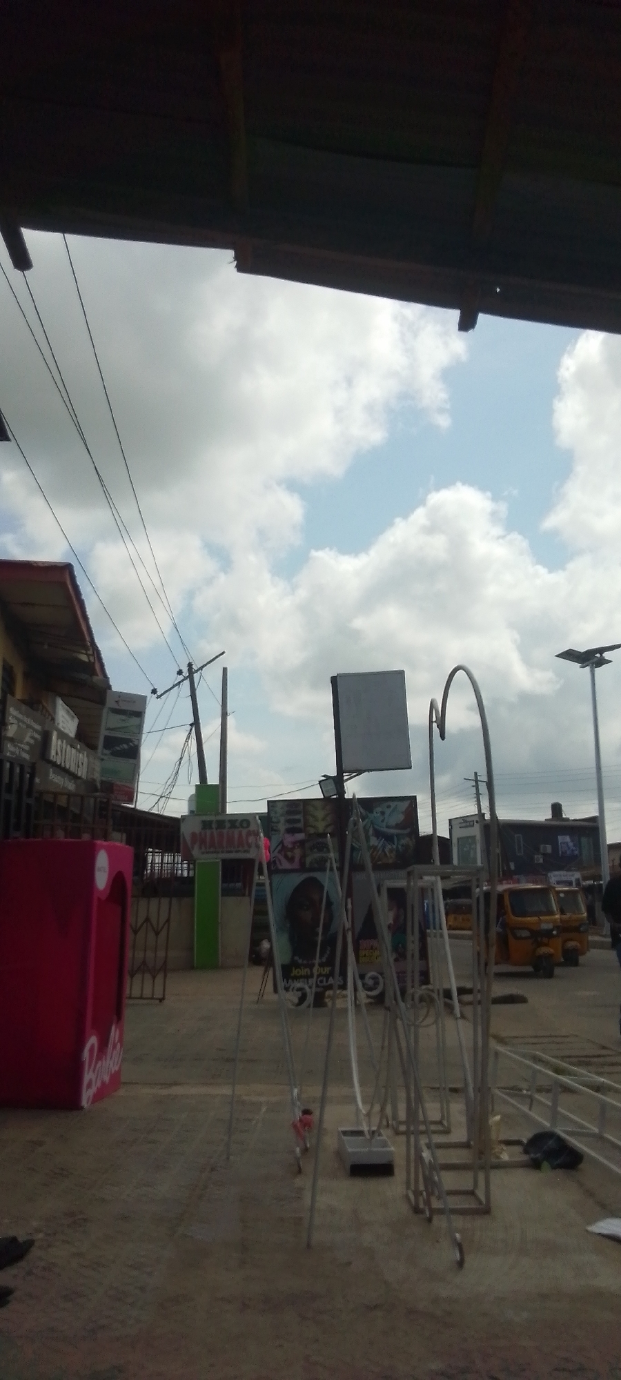 The image depicts a street scene in an urban area, likely in a developing country, based on the architecture and vehicles present. The view is partially obscured by a roof overhang in the foreground. In the midground, there's a pink box labeled "Barbie," possibly used for promotional purposes, standing next to a pharmacy, "NEXO PHARMACY." A structure of metal rods and arcs stands nearby, partially obscuring a sign advertising makeup classes. Beyond these elements, two yellow three-wheeled vehicles, resembling tuk-tuks or auto-rickshaws, are visible. Power lines run overhead. The sky is partly cloudy.