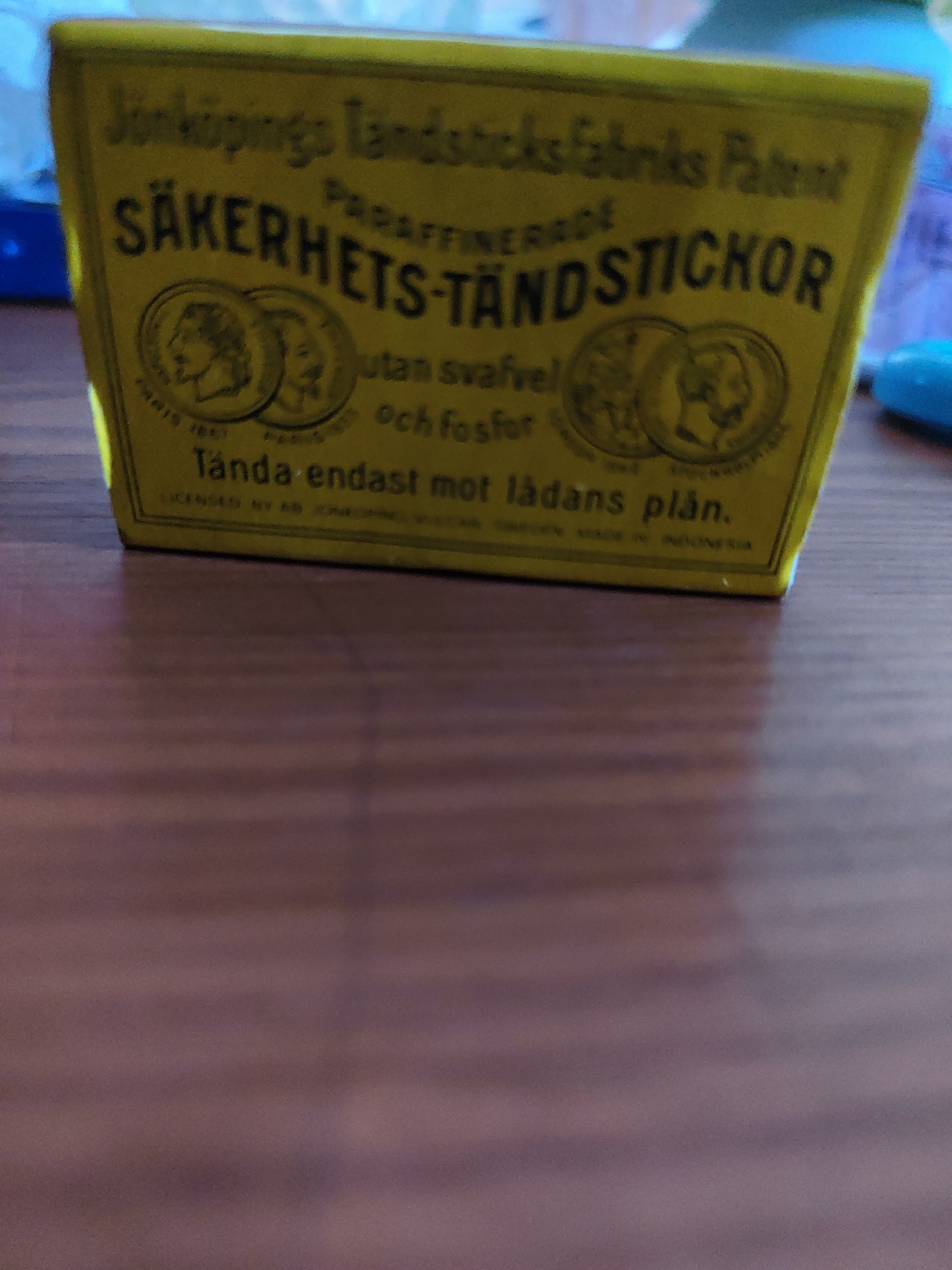 A yellow box of safety matches is resting on a wooden surface. The box has text and images printed on it. The text is in Swedish and appears to be a brand name and safety instructions. There are two circular emblems on the box, each containing a profile of a person. The wooden surface is a dark brown color and appears to be a table. The background is blurred and out of focus.