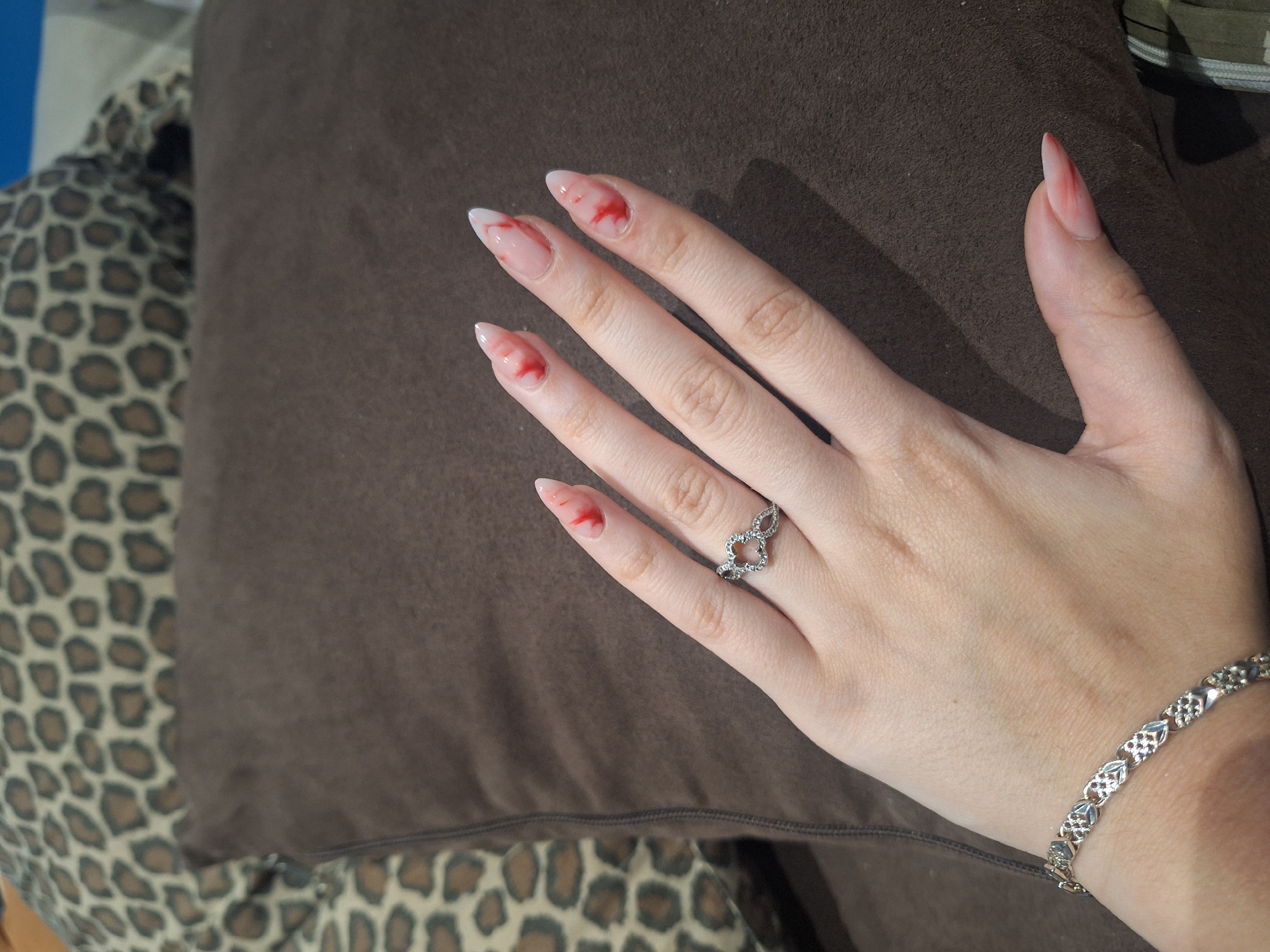 A close-up shot depicts a fair-skinned female hand with pointed, artificial nails, each adorned with a red, liquid-like substance near the tip. A silver ring with a heart-shaped design is on the ring finger. The wrist is adorned with a silver bracelet. The hand rests on a brown cushion with a smooth texture. The background includes a cushion or fabric with a leopard print pattern. There is no information available to determine the location in Sofia, Bulgaria.