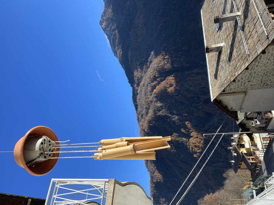 The image, taken in Dosso (Ciappanico), Italy, presents a vibrant, sunlit mountain vista. The perspective looks upwards and across a valley, with a clear blue sky dominating a significant portion of the frame.
In the foreground, a distinctive homemade wind chime hangs, suspended against the bright sky. It features an inverted, light brown terracotta pot at the top, from which multiple pieces of natural bamboo are hung by braided ropes, forming the chimes. Below the chime, parts of a white metal structure, possibly a railing or drying rack, and a section of a grey building facade are visible.
Beyond the immediate foreground, a large, steep mountain rises majestically. Its slopes are densely covered with trees, many of which display golden-brown foliage, indicating the autumn season. The sunlight highlights sections of the mountain, creating a play of light and shadow.
Towards the right side of the image, traditional multi-story buildings with stone walls and pitched roofs covered in dark, flat tiles are visible. These structures appear to be part of a village nestled at the base or on the lower slopes of the mountain, further down in the valley.
The scene depicts a clear, sunny day, likely during midday or early afternoon, given the bright illumination. There are no people visible, nor any specific activities taking place, creating a serene and picturesque landscape. No text is visible in the image.