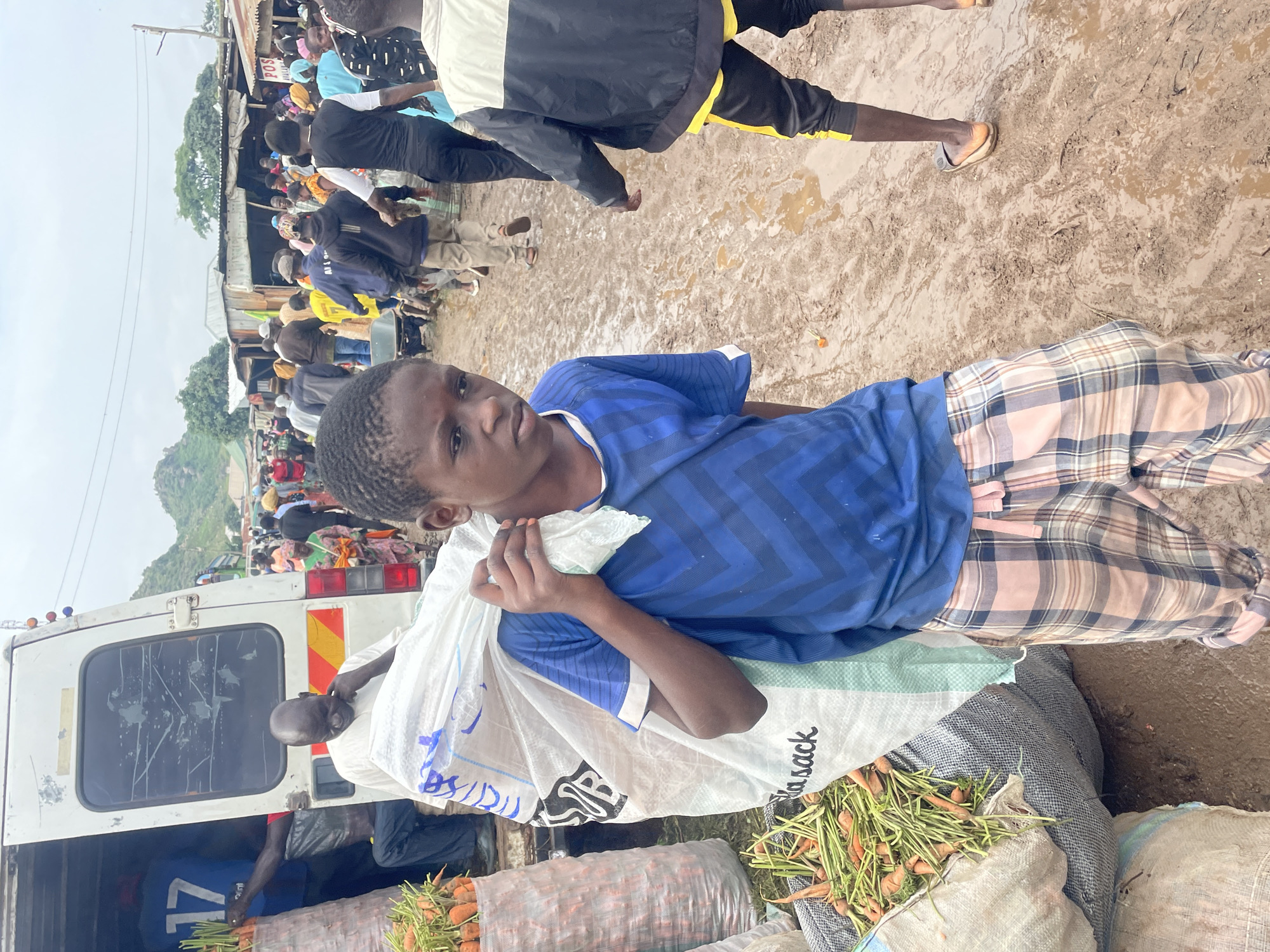 In Gura Top, Nigeria, a young boy, likely under 16, carries a large, white sack of produce over his right shoulder. He wears a blue, chevron-patterned t-shirt and plaid trousers. To his left, a white van with an open rear door is parked. Sacks of carrots are visible near the van, one large gray sack filled with carrots sits to the boy's left. The ground is muddy, indicating recent rainfall. In the background, a bustling market scene unfolds with numerous people, temporary structures, and stalls. The sky is overcast.