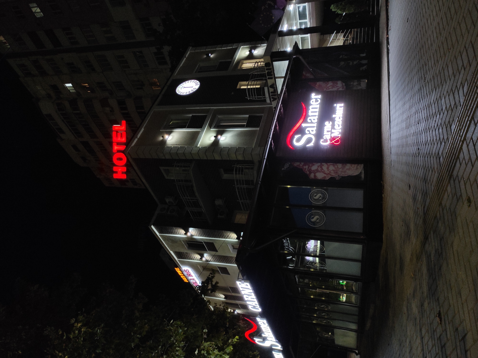 A low-angle, exterior shot at night captures a modern building illuminated by artificial lights. A prominent red neon sign reading "HOTEL" is mounted on the upper facade. Below the hotel sign, another neon sign, this one in white and red, displays "Salamualaikum CARNE & Mezeluri" which translates to "Salamer Meat & Sausages". The building appears to be a hotel and restaurant. The foreground is paved with interlocking bricks. A tall apartment building is visible in the background, also lit from within, suggesting it is occupied. The scene is set in an urban environment.