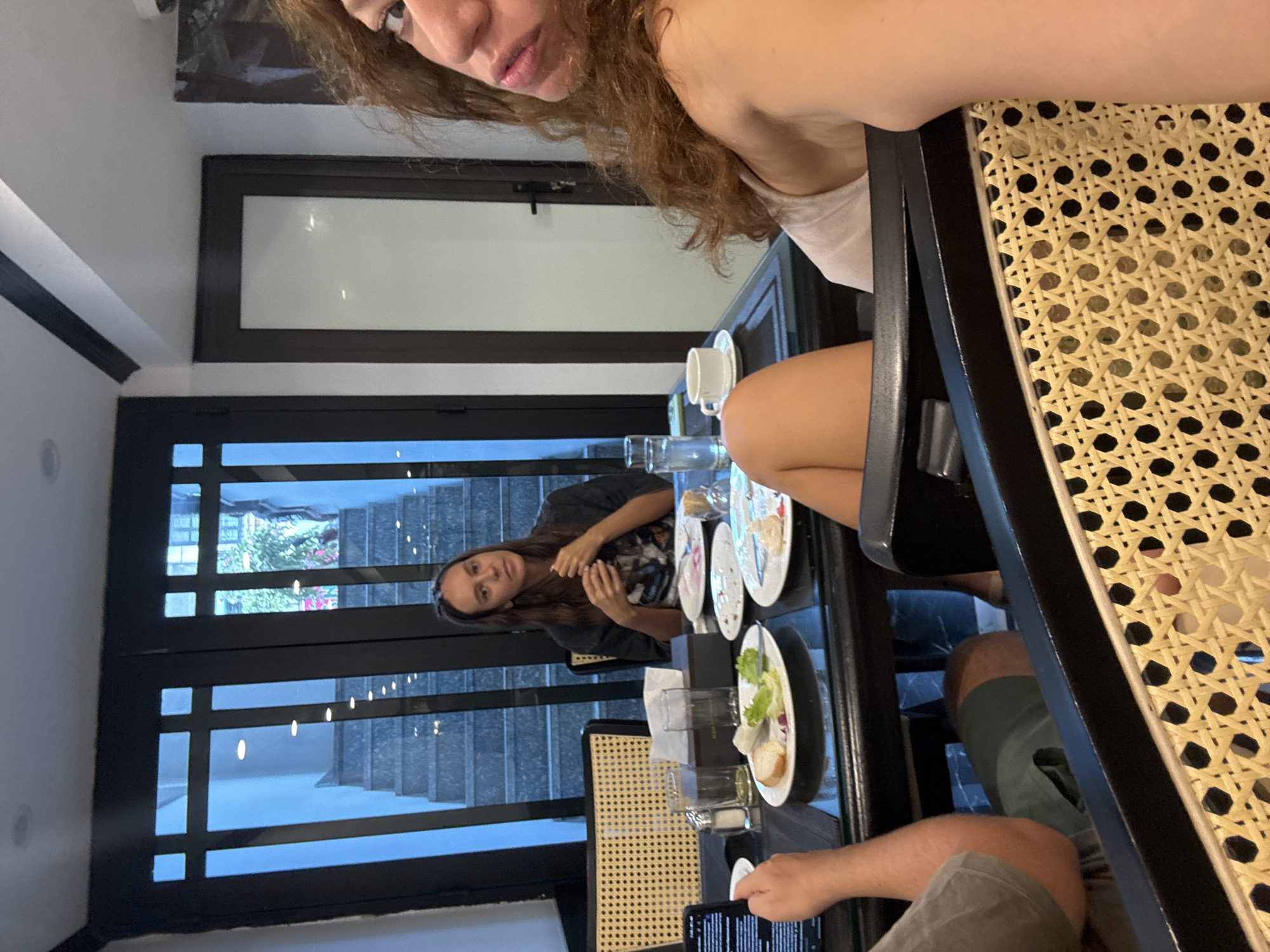 The image captures a restaurant scene in Hà Nội, Vietnam, as viewed by an individual at the table. The restaurant has a modern architectural design with large glass panel windows overlooking a stairwell. The perspective is from someone seated at a dark wooden table laden with dishes, glasses, and cutlery, suggesting a meal has been eaten or is in progress. A second woman is seated across the table, engaged with eating. The presence of a smartphone held by a man, partly visible in the foreground, suggests photography or browsing is occurring. The scene is brightly lit, likely with natural light coming through the windows. The focus is on the social interaction, the food setting, and the indoor environment, with the urban landscape visible in the background.