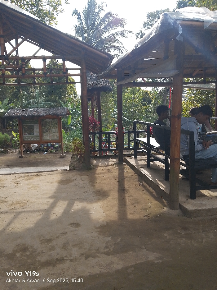 The image depicts an outdoor recreational area in Sukadana, Indonesia, under clear daylight conditions with visible shadows indicating sunlight. The scene is dominated by two rustic, open-sided shelters or gazebos constructed from bamboo and wood. The roofs of both structures appear to be thatched with organic material, with the rightmost shelter additionally showing clear plastic sheeting on its roof.
On the left side of the frame, a wooden information board with a thatched roof stands upright, displaying text and images. The ground in the foreground is unpaved, consisting of packed dirt with some uneven textures and darker patches possibly indicating moisture. Lush green tropical foliage, including large palm trees and banana plants, forms the background, suggesting a natural or park-like environment. Red flowering plants are visible near the bases of the shelters.
Inside the rightmost shelter, three young males are seated on a dark-colored bench. The individual on the far left of the bench is partially obscured and wears a dark shirt. The middle person wears a light-colored, long-sleeved top and is looking downwards, possibly at a personal device or item. The person on the far right is also looking down, partially obscured by the middle individual, wearing a dark top. No direct interaction between the seated individuals is discernible; their postures suggest individual engagement or passive resting. The shelter on the right features dark-colored railings.