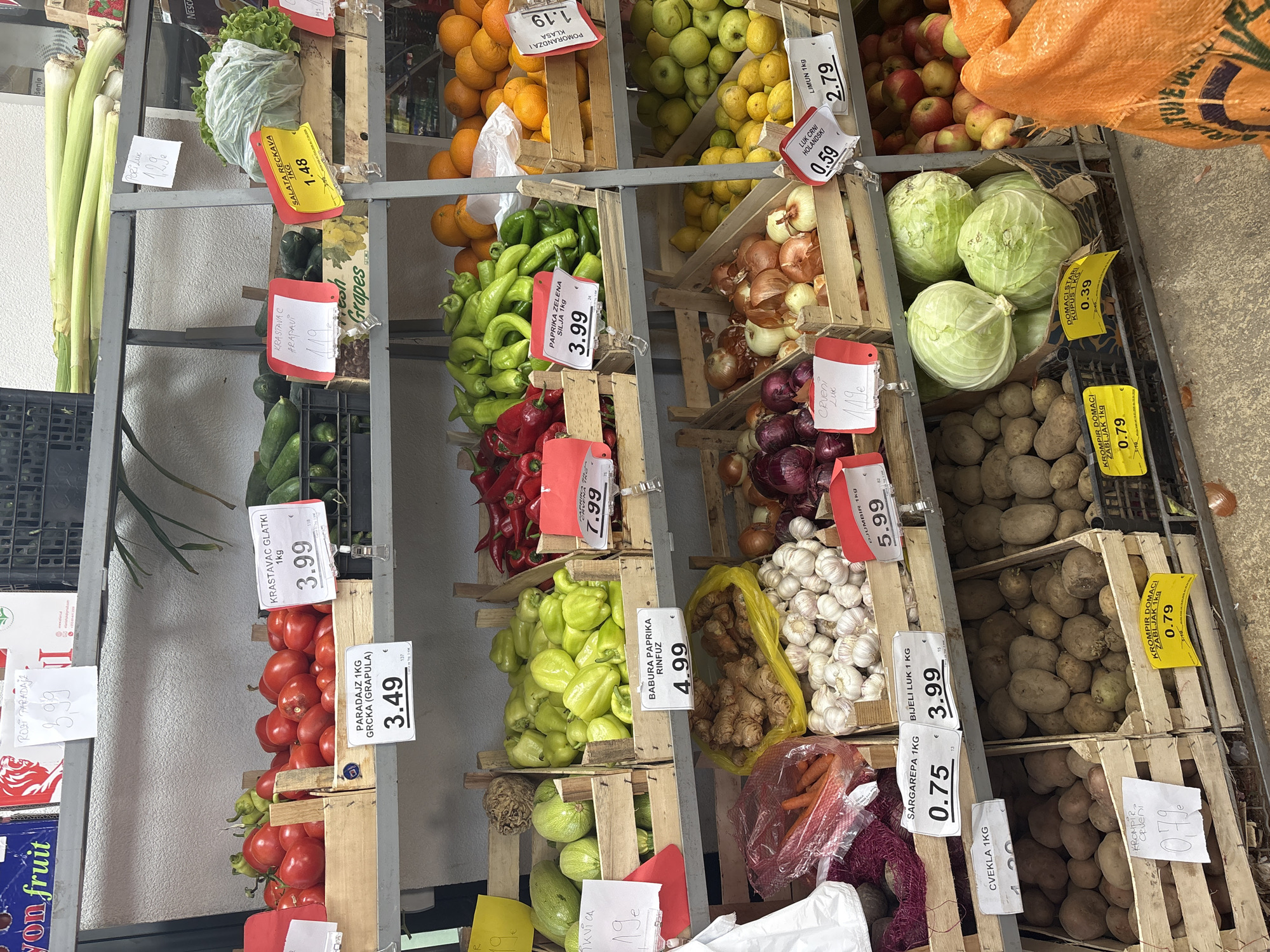 This image captures a vibrant market stall or greengrocer's display, showcasing a variety of fresh produce neatly arranged in wooden and plastic crates on metal shelving. The bright, natural lighting suggests it is daytime.
The subjects include an abundance of fruits and vegetables such as spring onions/leeks, crisp lettuce, bright oranges, green and yellow apples, lemons, green pointy peppers, red bell peppers, yellow and red onions, heads of garlic, ginger roots, carrots, beets, light green cabbages, red and round tomatoes, cucumbers (both smooth and "arapavi" or ridged types), and various potatoes. An orange sack partially covers some apples at the top right, while other produce like carrots and beets are in clear plastic bags.
The activity depicted is the sale of fresh produce at a market. The items are organized for display, with price tags clearly visible for each product.
Notable details include the extensive use of price tags, many handwritten, indicating prices in Euros (€). The text on these tags is predominantly in Montenegrin or a similar South Slavic language, with product names like "PARADAJZ 1KG GRCKA (GRAPULA)" (tomatoes) for 3.49€, "PAPRIKA ZELENA SILJA 1kg" (green pointy peppers) for 3.99€, "KROMPIR DOMACI ZABLJAK 1KG" (domestic potatoes from Žabljak) for 0.79€, and "LIMUN 1kg" (lemons) for 2.79€. Other visible text includes "SALATA RECKAVA 1KG" (lettuce) for 1.48€, "BIJELI LUK 1 KG" (white garlic) for 3.99€, and "CVEKLA 1KG" (beets) for 0.75€. The currency and language, consistent with the location context of Bar, Montenegro, suggest a local market environment.