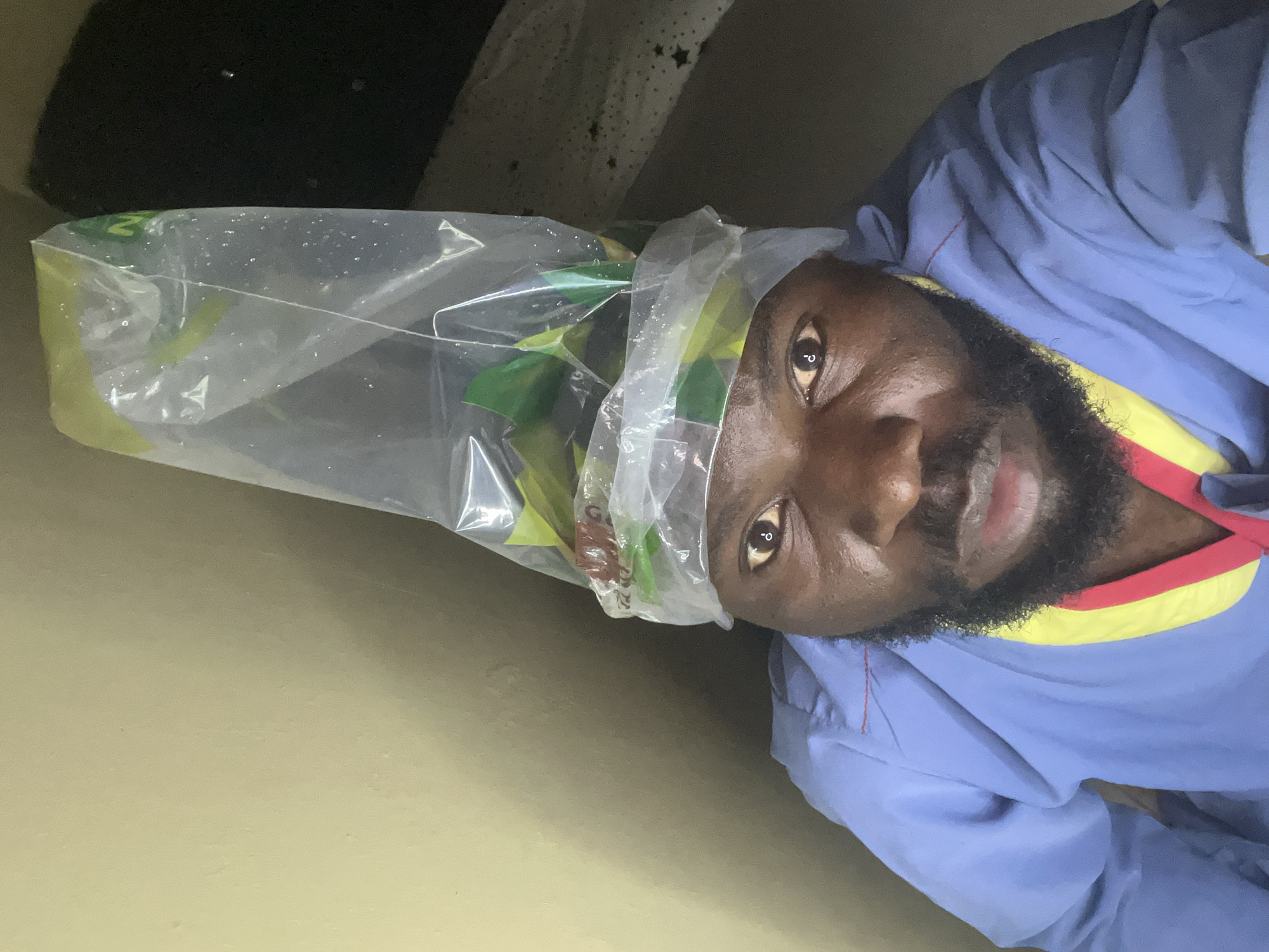 A man with dark skin and a beard is depicted in a medium close-up, looking directly at the camera. He is wearing a light blue top with a red and yellow trim around the collar, reminiscent of scrubs or a work uniform. A transparent plastic bag is placed over his head, appearing to contain green, leafy items or produce, along with possibly other colored objects. The inside of the plastic bag shows condensation or small water droplets.
The man's expression is calm and neutral, with a slight, subtle smile. His eyes are open and engaged with the viewer. The background consists of a plain, light beige or tan wall or surface, with a darker, shadowed area in the upper left corner, suggesting an indoor or shaded environment. The lighting is somewhat dim, highlighting the man's face and the bag.
There is some barely discernible and illegible text visible through the plastic bag, possibly on the green items inside and faintly on the bag itself (e.g., a distorted 'Z' or similar characters). No specific location cues are visible in the image, but the context indicates the setting is Gura Top, Nigeria. The activity appears to be a posed photograph, possibly playful or for documentation, with the man deliberately wearing the bag on his head.