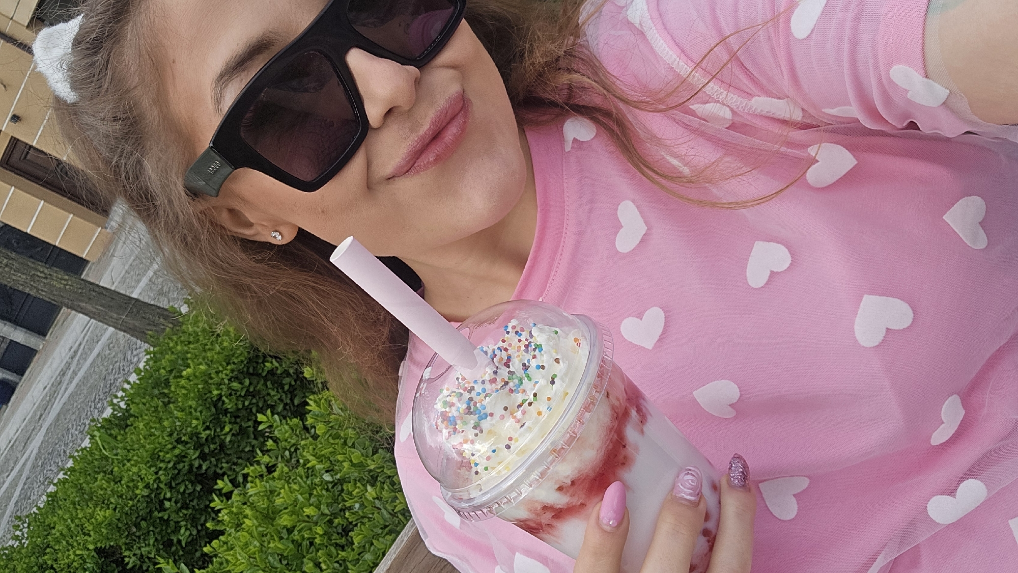 A young woman poses in a selfie, possibly outdoors, holding a large clear plastic cup filled with a strawberry milkshake, topped with whipped cream and colorful sprinkles. She is wearing a pink top with white hearts and black sunglasses. Her nails are painted pink. The setting appears to be an urban area with green shrubs visible in the background, possibly in Elhovo, Bulgaria. The photograph is taken at an angle.