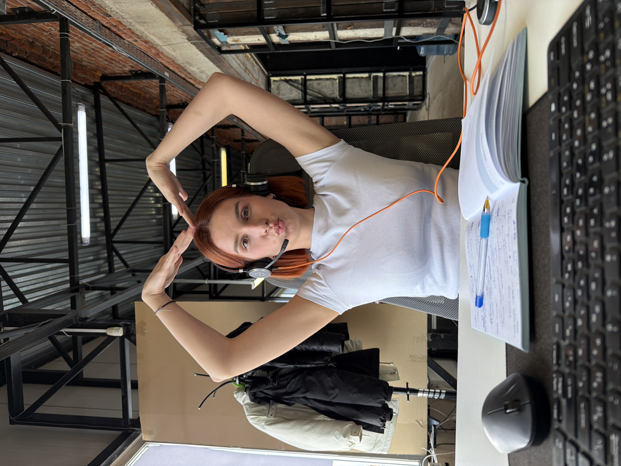 The image depicts a female subject in an office or workspace located in Chaikovsky, Russia. The subject, with reddish-brown hair, is seated in a grey office chair, facing the camera. She is wearing a light-colored ribbed short-sleeved t-shirt and a headset with an orange cord. Her arms are raised, with her hands forming a heart shape above her head, and her lips are pursed in a pout. A silver-colored bracelet is visible on her left wrist.
On the desk in the foreground, from left to right, are a dark grey computer mouse, an open notebook with blue ink writing and a blue pen resting on it, and a partial view of a black computer keyboard. The orange headset cord runs across the desk.
The background features an exposed industrial-style ceiling with corrugated metal, black support beams, and fluorescent tube lighting fixtures. To the left, a window provides natural light, and a coat rack holds a cream-colored puffer jacket and a black jacket, with a large cardboard panel behind it. To the right, an exposed red brick wall is visible alongside industrial-style shelving units or structures.