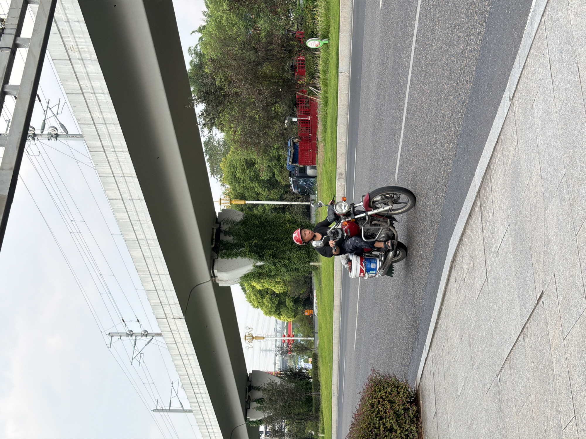An individual wearing a red helmet and a black t-shirt rides a red motorcycle with a white bucket and blue container attached to the rear. The motorcycle is on an asphalt road, with a gray paved sidewalk to its left and a green median strip to its right. In the background, a large concrete overpass for an elevated railway system dominates the scene, with electrical wires visible above it. Lush green trees and some structures, including a red fenced area and a blue vehicle, are situated beyond the median. The sky is overcast. The image appears to be taken in the city of Jiangjin, China.