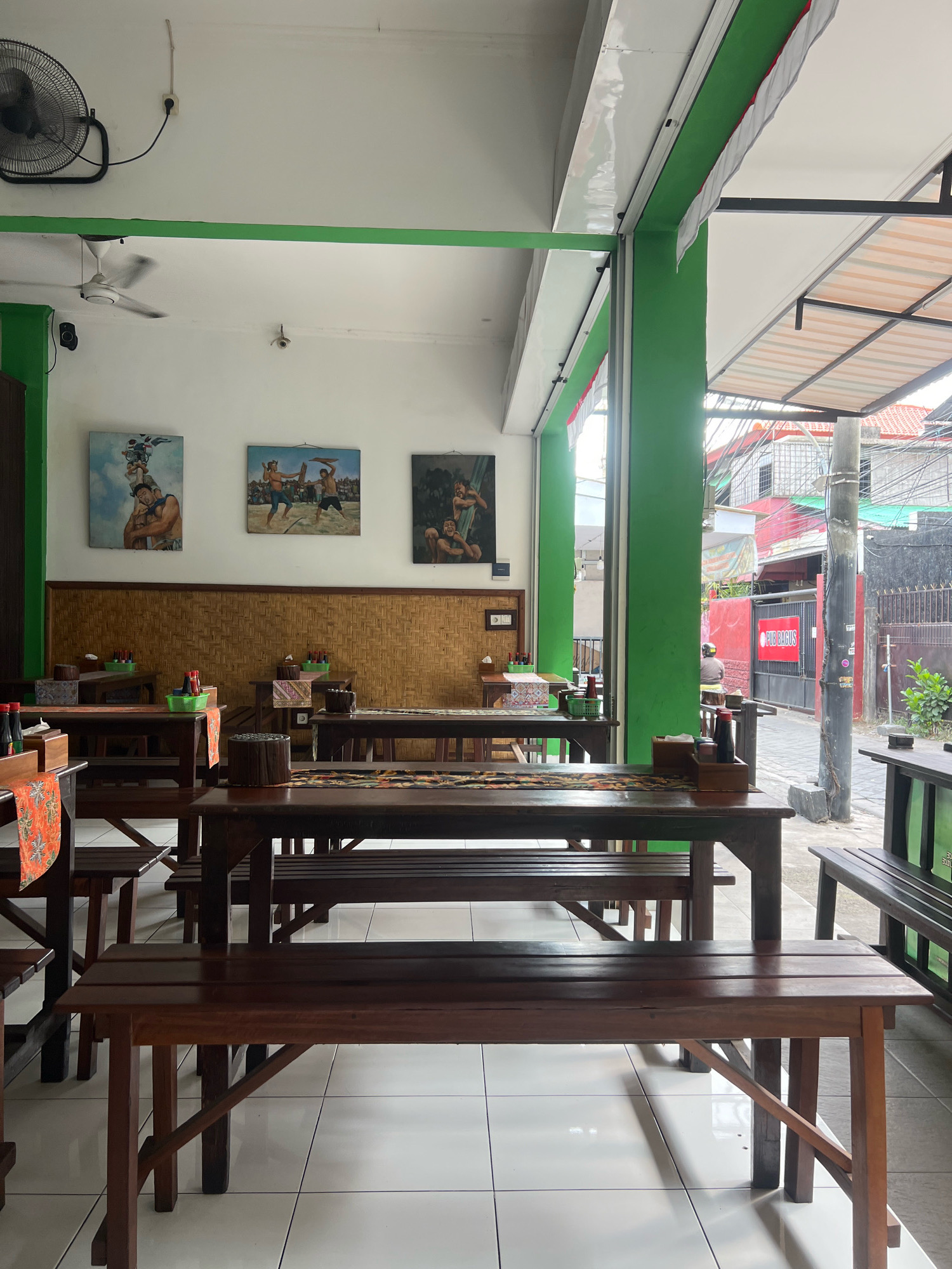 The image depicts the interior of a restaurant, likely in Kuta, Indonesia, with a focus on the seating area. The restaurant features numerous wooden tables and benches, arranged in rows. The walls are painted white and green, with decorative paintings of local scenes on the upper wall. Woven bamboo panels form a lower section of the walls, behind the tables. Each table is set with condiments in bottles, small green baskets, and decorative placemats. The floor is tiled with white squares. Natural light streams in from an open doorway or window on the right side of the image, revealing a street scene with buildings and a power pole outside. A ceiling fan is visible on the upper left, and a wall-mounted fan is on the upper left wall. The overall impression is of a casual, open-air dining establishment.