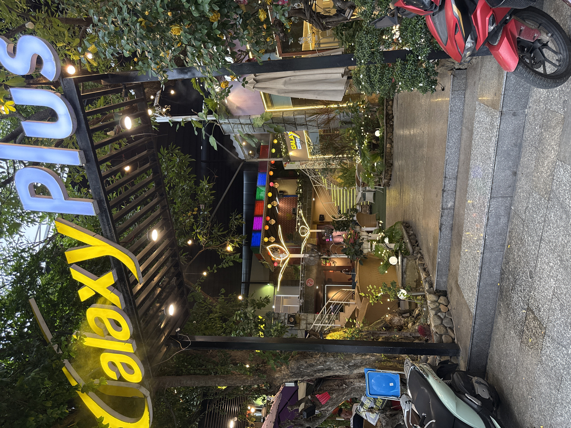 The image captures the entrance of "Galaxy Plus Coffee" in Nha Trang, Vietnam. The establishment features a prominent sign with large, illuminated lettering spelling out "Galaxy Plus" in yellow and blue. The entrance is covered by a wooden trellis adorned with small, warm-toned string lights, creating a festive atmosphere. Lush green trees and foliage surround the entrance, with yellow flowers blooming on one side.
The pathway leading into the coffee shop is paved with grey stone tiles, and there are several steps leading up to the main entrance area. The interior of the coffee shop is visible through the entrance, showcasing a well-lit space with stairs, seating areas with wicker furniture, and more decorative lighting. To the left of the entrance, a light-colored scooter is parked on the pavement, and to the right, a red motorcycle is partially visible. A man is seated on a chair to the left, near a tree. The overall scene suggests a welcoming and vibrant ambiance for a coffee shop.