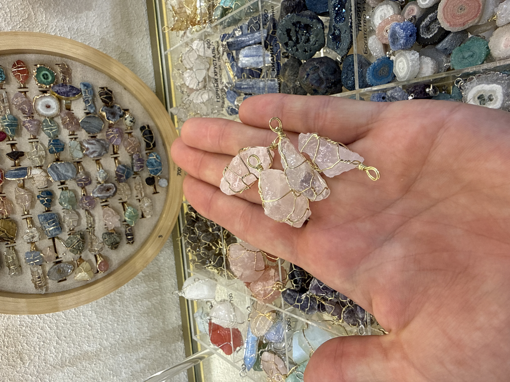 A close-up shot shows a person's hand holding a cluster of rose quartz crystals wrapped in gold wire. The crystals are light pink and appear to be raw, unpolished pieces. In the background, a display of various crystals and semi-precious stones is visible. These include rings with colorful stones arranged in a wooden circular display on the left, and a tiered display of pendants and raw stones in clear plastic containers on the right. Some of the stones appear to be geode slices with intricate patterns and vibrant colors like blue, green, pink, and white. Labels with Russian text and numbers like "400," "500," "700," and "1000" are visible on the displays, suggesting prices or inventory codes. The setting appears to be a shop or market stall selling crystals and jewelry. The lighting is bright, indicating it might be daytime indoors or under bright artificial lights. The overall mood is one of commercial display and the appreciation of natural stones.