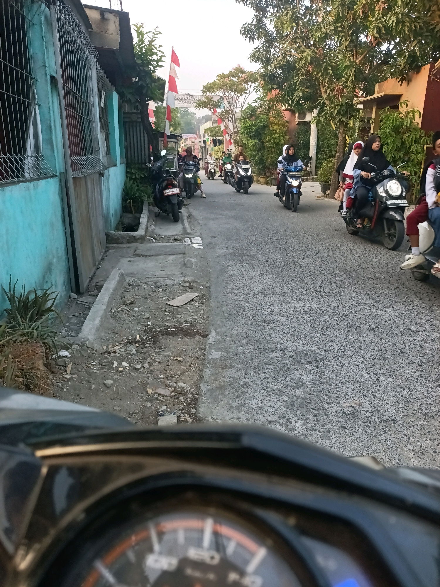 The image depicts a street scene in Wanasari, Indonesia, as indicated by a banner that reads "SELAMAT JALAN RT 02 RW 08". The perspective is from the viewpoint of someone riding a motorcycle, with the motorcycle's dashboard visible in the foreground, slightly out of focus.
The street is paved with asphalt and is lined with houses and vegetation. On the left side of the street, a turquoise-colored building with metal bars over its windows is visible. A narrow, somewhat dilapidated sidewalk runs alongside the building, with exposed earth, stones, and debris. Several potted plants and a larger plant with spiky leaves are present near the sidewalk.
The right side of the street features trees and residential buildings, one with an orange-brown facade. A line of motorcycles is traveling down the street. Several individuals are riding on these motorcycles, most of whom appear to be wearing headscarves, suggesting a predominantly Muslim population. One motorcycle in the mid-ground has two young girls on it, one wearing a red and white hijab and a school uniform. Another motorcycle further ahead has a rider wearing a blue hijab. The motorcycles are of various models and colors.
Red and white Indonesian flags are prominently displayed, attached to poles along the street, indicating a festive or commemorative occasion. The lighting suggests it is daytime, possibly late afternoon, with a soft, diffused light. The overall atmosphere appears to be one of everyday movement and community activity within a residential urban environment.