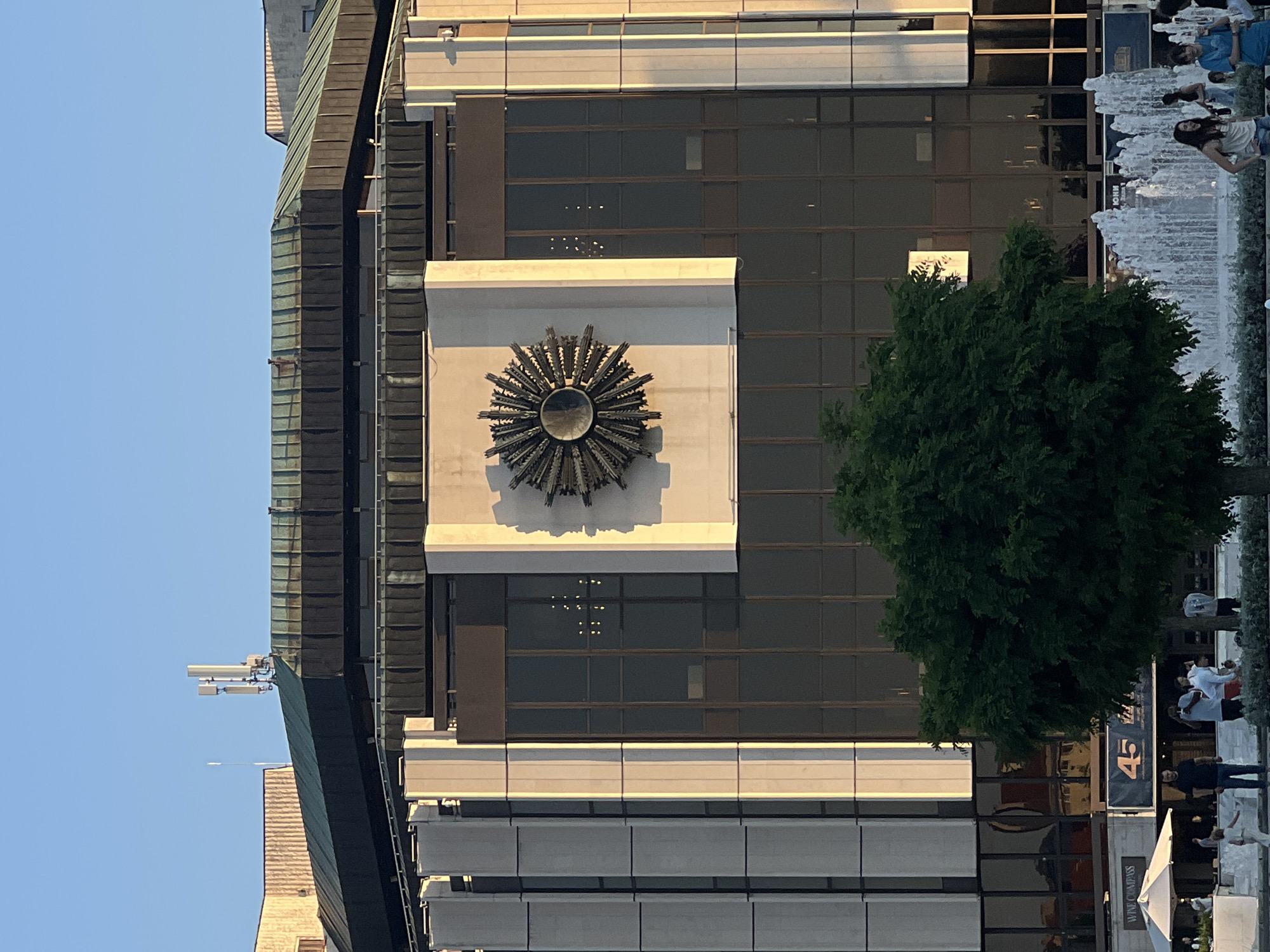 The image shows a large, multi-story building with a dark, angled roof. A prominent, starburst-shaped bronze sculpture is mounted on the upper facade of the building. The building has a glass exterior with vertical white columns on either side of the sculpture. Below the sculpture, a large tree obscures the lower portion of the building. In the foreground, a fountain with multiple water jets is visible, with several people standing near it. Some of these individuals are looking towards the camera, while others are engaged in conversation. A white umbrella shading an outdoor seating area and a banner with the number "45" are also visible in the lower left and center of the image, respectively. The sky is clear and blue, and the sunlight casts a warm glow on the building.