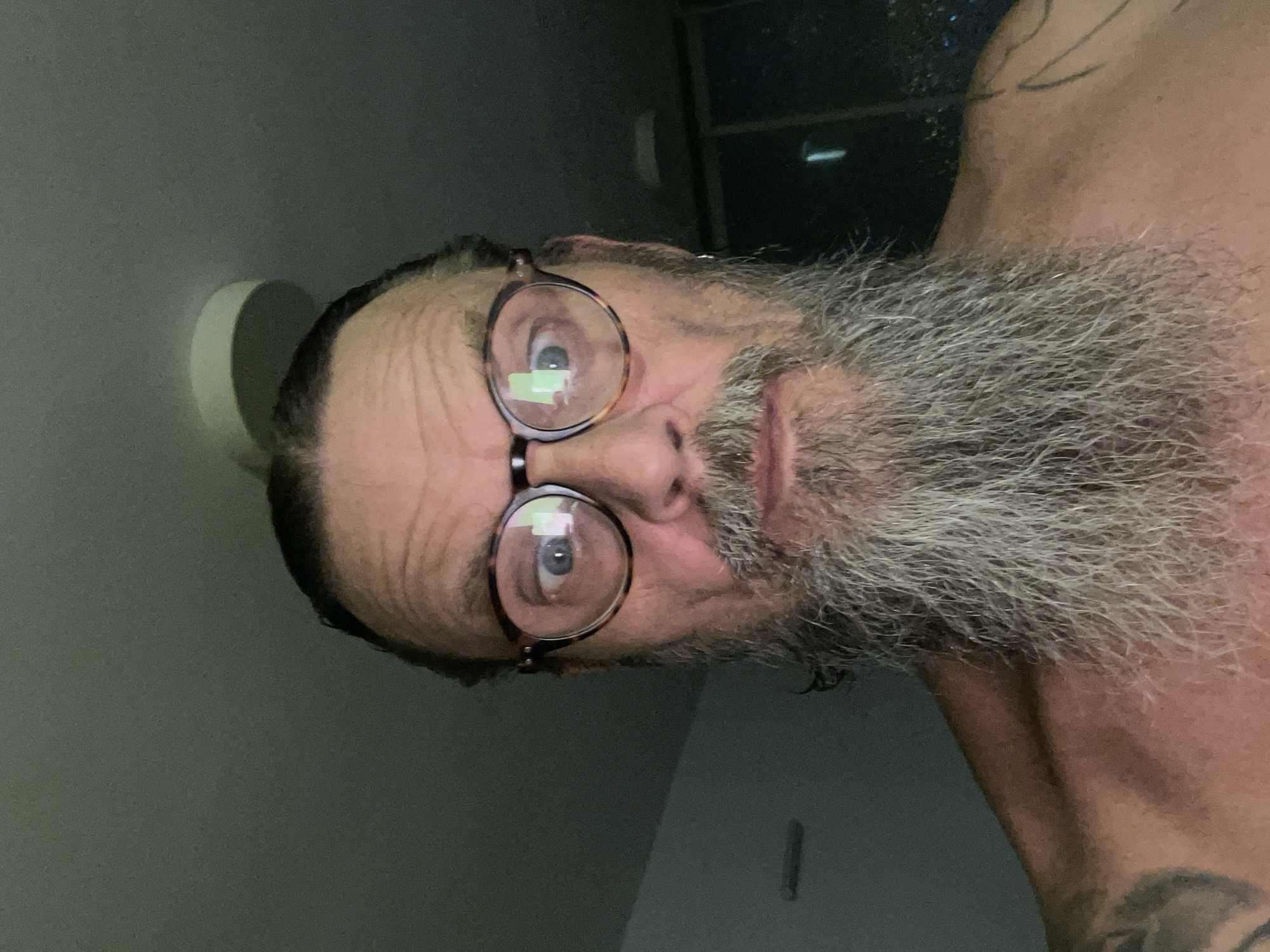 A close-up, head-and-shoulders selfie shows a shirtless man with a full, graying beard and long, dark hair pulled back. He is wearing tortoise-shell glasses, and his eyes are looking in slightly different directions. The background is dark and appears to be an indoor environment with a light fixture visible on the ceiling. There are some faint reflections in the man's glasses. A portion of a tattoo is visible on his left shoulder. The image was taken in the city of Netanya, Israel.