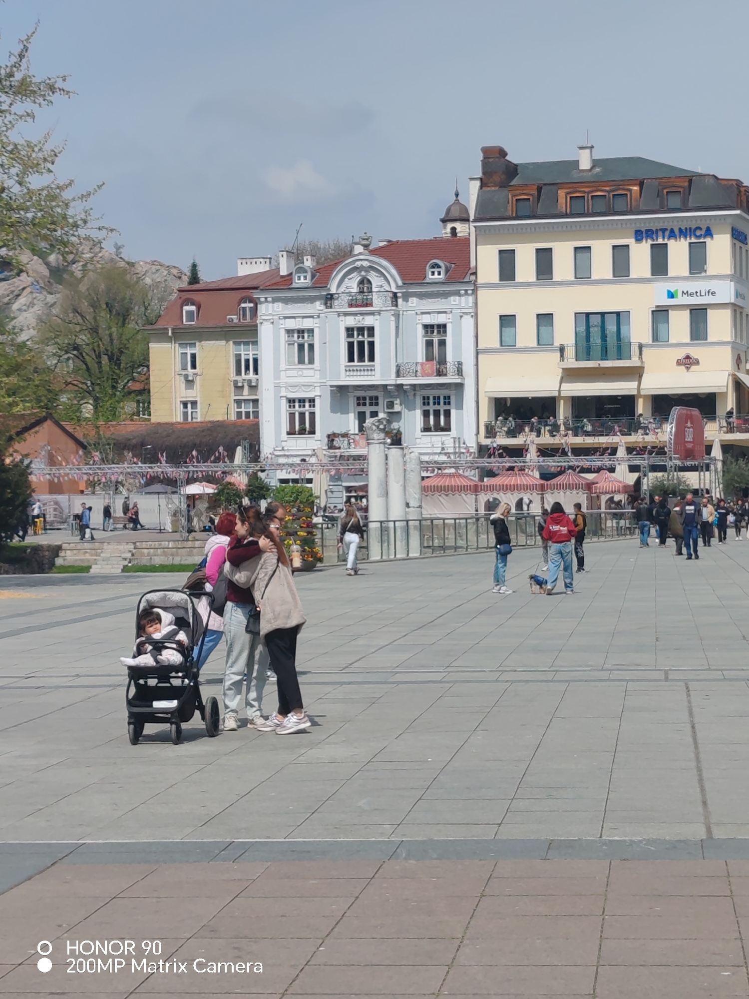 The image captures a wide shot of a public square in Plovdiv, Bulgaria, possibly the Central Square. Prominent buildings frame the scene, including one with "BRITANICA" and "MetLife" signage. A central fountain area with tents and decorative elements is visible in the middle ground. Several groups of people are scattered across the square. In the foreground, a baby is seated in a stroller, near a group of three people interacting. Others are strolling or standing around in the square. The square is paved with light grey stones. In the background, there are buildings and mountains. The weather is sunny.
