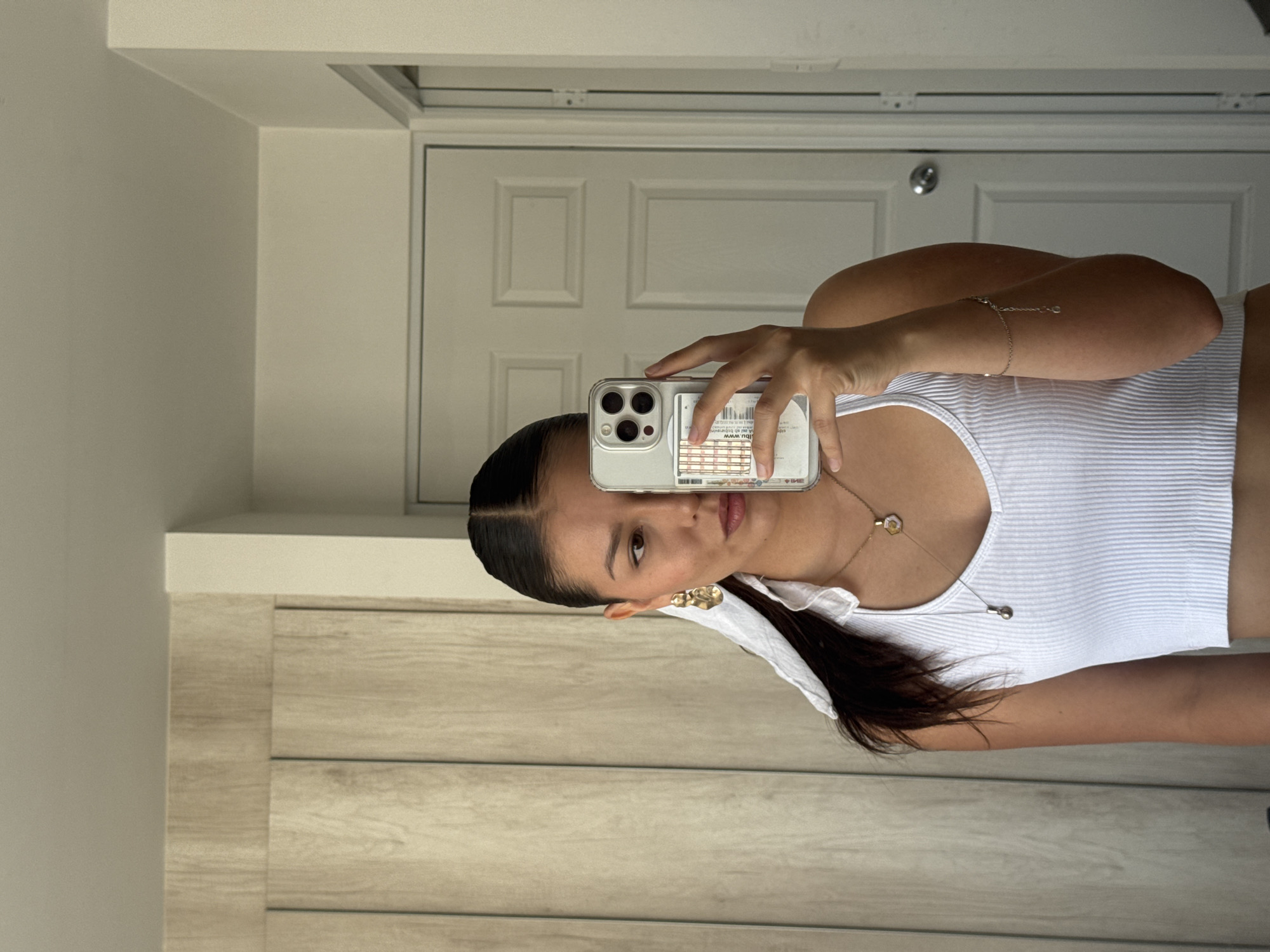 A woman in a white ribbed tank top holds up her phone in front of a mirror to take a selfie. Her hair is dark and pulled back at the top. She wears a necklace and earrings. The background includes a white door with three panels and white walls. The location cannot be determined to be San Francisco Ocotlán, Mexico from the image.