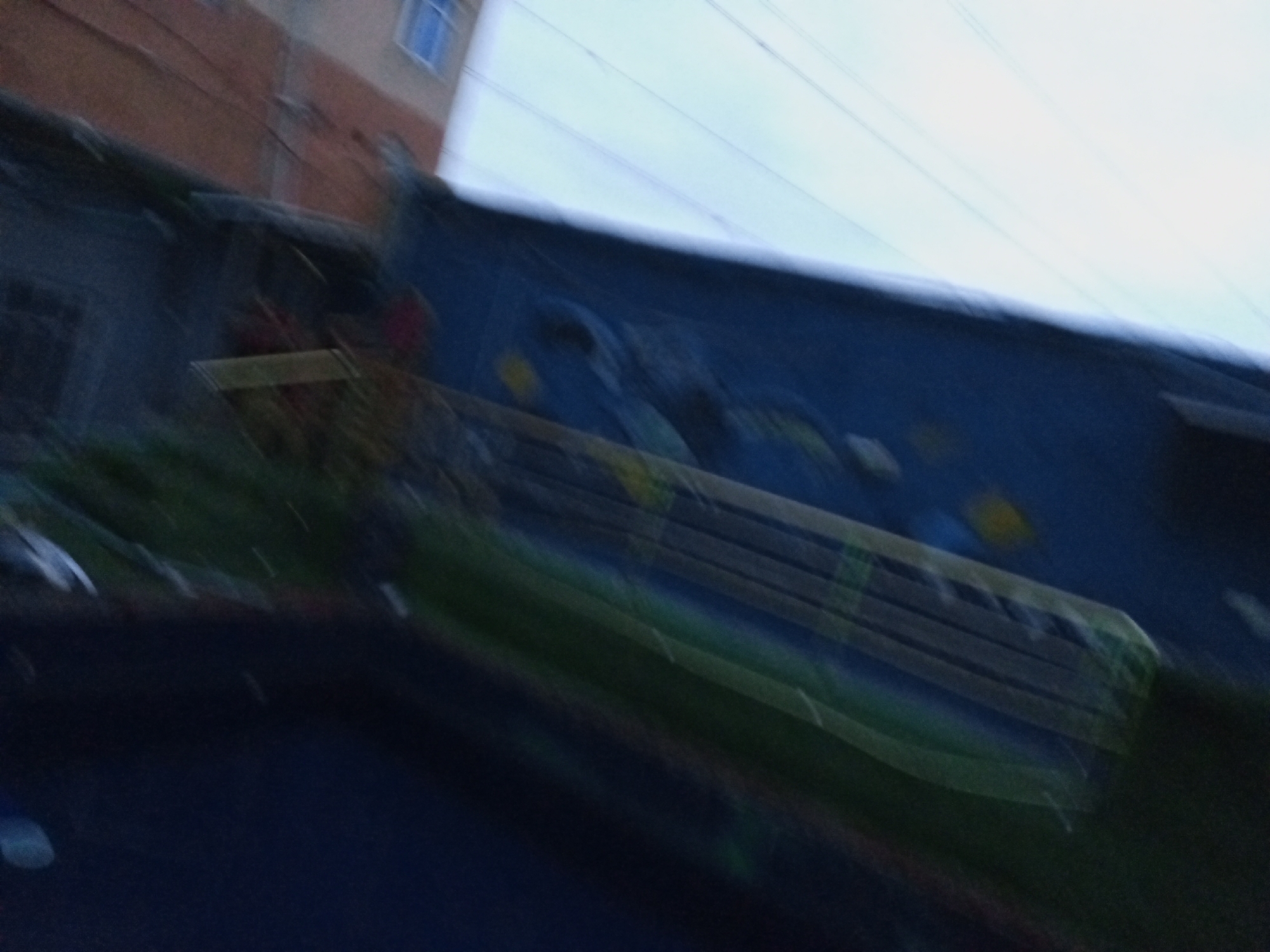 The image captures a blurred view of a building and what appears to be a mural or painted wall in the city of Ado, Nigeria. The building on the left is partially visible, with a reddish-brown upper section and a lighter section below. A blue window with a grid pattern is discernible in the upper part of the building. The main focus of the image is a large, dark blue surface, possibly a wall or the side of a structure, adorned with a mural. The mural is abstract and colorful, with streaks of yellow and green visible. There is a suggestion of a long, horizontal element with lines, possibly a part of the mural design or an object placed in front of it. The foreground is dark and blurry, with hints of green foliage or vegetation. The overall impression is one of an outdoor scene, likely in an urban setting, with the motion blur suggesting movement during the capture of the image.