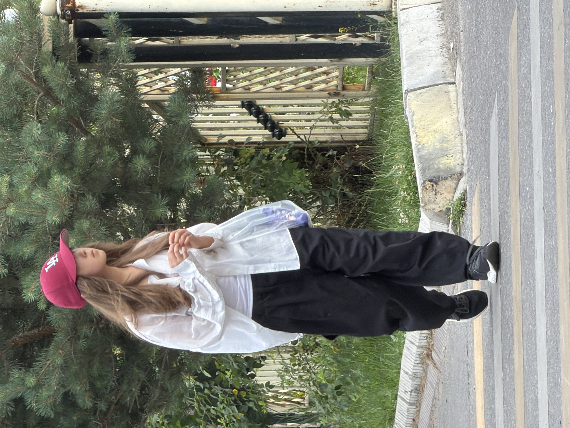A young female walks across a pedestrian crossing in an outdoor, urban environment. She is wearing a red baseball cap, a white button-down shirt over a white t-shirt, and loose-fitting black pants. She carries a clear plastic bag in her right hand. The background features green foliage, a metal fence, and a pine tree. The ground is asphalt with yellow and white painted lines indicating the pedestrian crossing. The lighting suggests it is daytime.