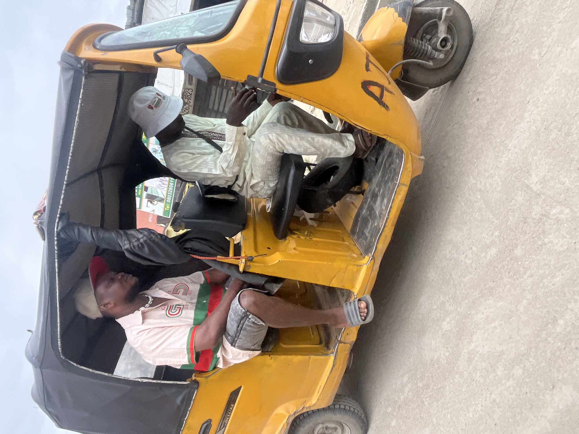 A side view, medium shot, captures two men inside a bright yellow auto-rickshaw. The rickshaw, a common mode of transportation in Ado, Nigeria, features a black canopy for shade. The man in the foreground, seated on the left, wears a light pink short-sleeved shirt with red and green horizontal stripes across the chest, grey denim shorts, and grey flip-flops. He has a tan baseball cap with a red brim and is looking to his right. Around his neck is a shell necklace. The man in the driver's seat, on the right, is wearing a light-colored traditional outfit and a white bucket hat with a Nigerian flag emblem. He is looking down and appears to be operating the vehicle. The rickshaw's exterior has the letters "AT" visible in black paint. The vehicle is on a paved road, with a concrete surface in the foreground. In the background, buildings and possibly other vehicles are visible under an overcast sky.