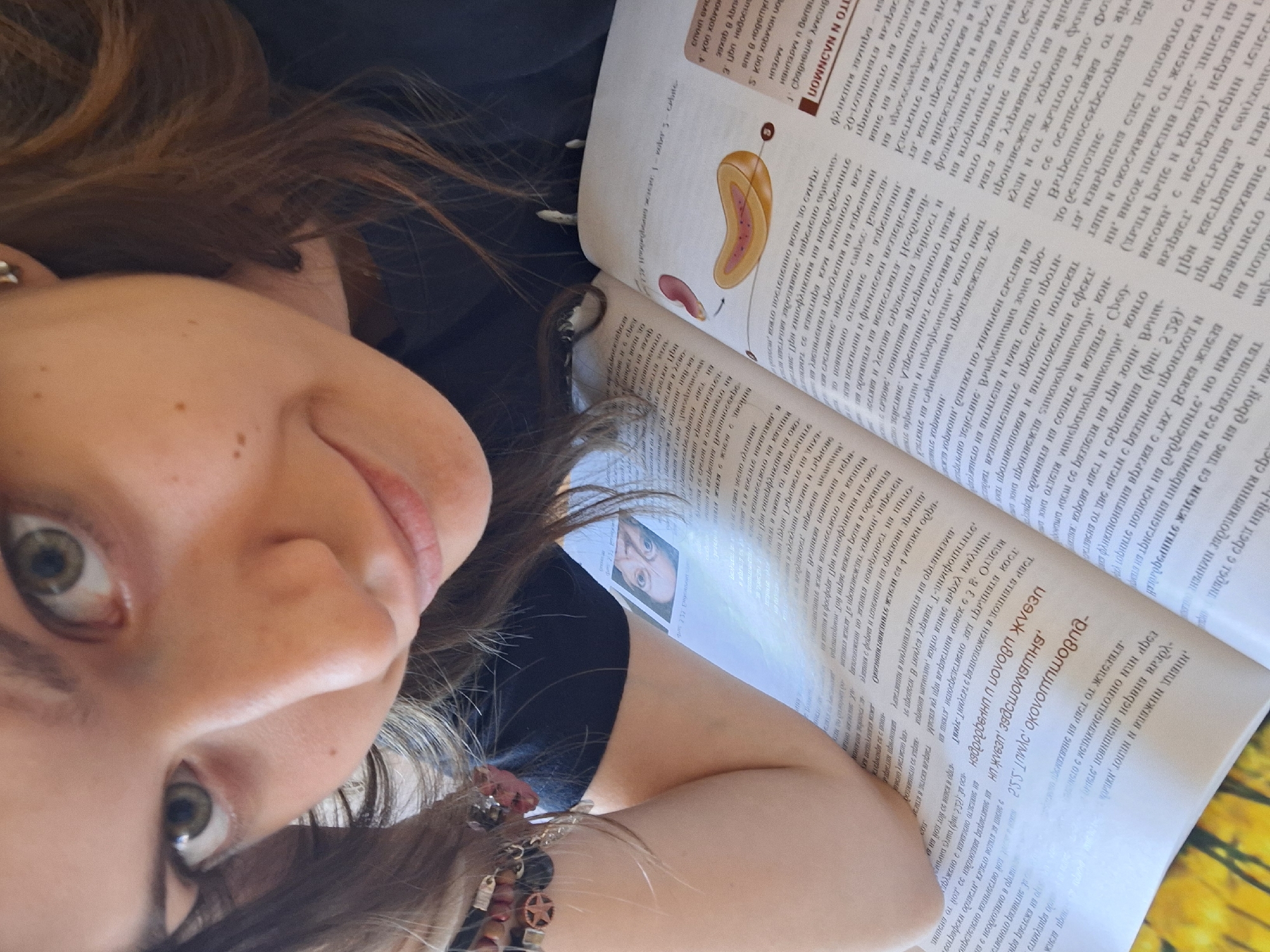 A young woman is pictured in a selfie, lying down and facing the camera. She has fair skin, hazel eyes, and light brown hair, a portion of which is visible on the left side of the frame. She is wearing a black shirt and a bracelet on her left wrist. The woman is lying next to an open textbook, which is filled with Bulgarian text and diagrams. The pages of the book take up a significant portion of the frame and appear to contain scientific or medical information. Based on the image data provided and the fact that the text is Bulgarian, it cannot be reliably determined where this photo was taken, especially not in the city of Panagyurishte.