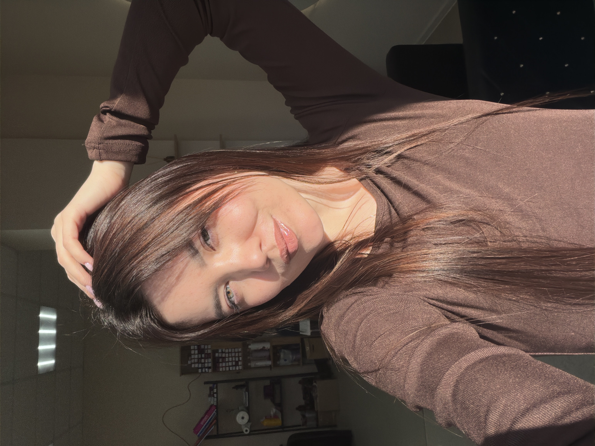 A young woman with long, brown hair and light skin is posing for a selfie in what appears to be an indoor environment. She is wearing a brown long-sleeved shirt. Her right hand is positioned above her head, with sunlight illuminating her face and hair. In the background, there's a bookshelf and a dark chair. The location is suggested to be Chaikovsky, Russia.