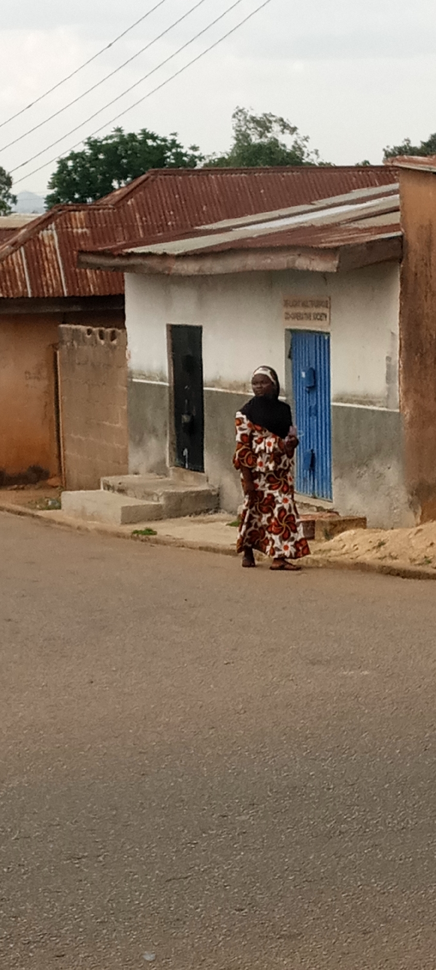 A woman in a long patterned dress and a black headscarf stands on a paved road in front of a small, simple building. The building has a corrugated metal roof that is reddish-brown, and its walls are light gray. There are two doorways, one with a dark, possibly metal door, and another with a blue corrugated metal door. A small sign is visible above the blue door. A short flight of concrete steps leads up to the doorways. To the left of the building is another building with similar construction and rust-colored walls. Power lines are visible against the overcast sky. The location appears to be an urban area.